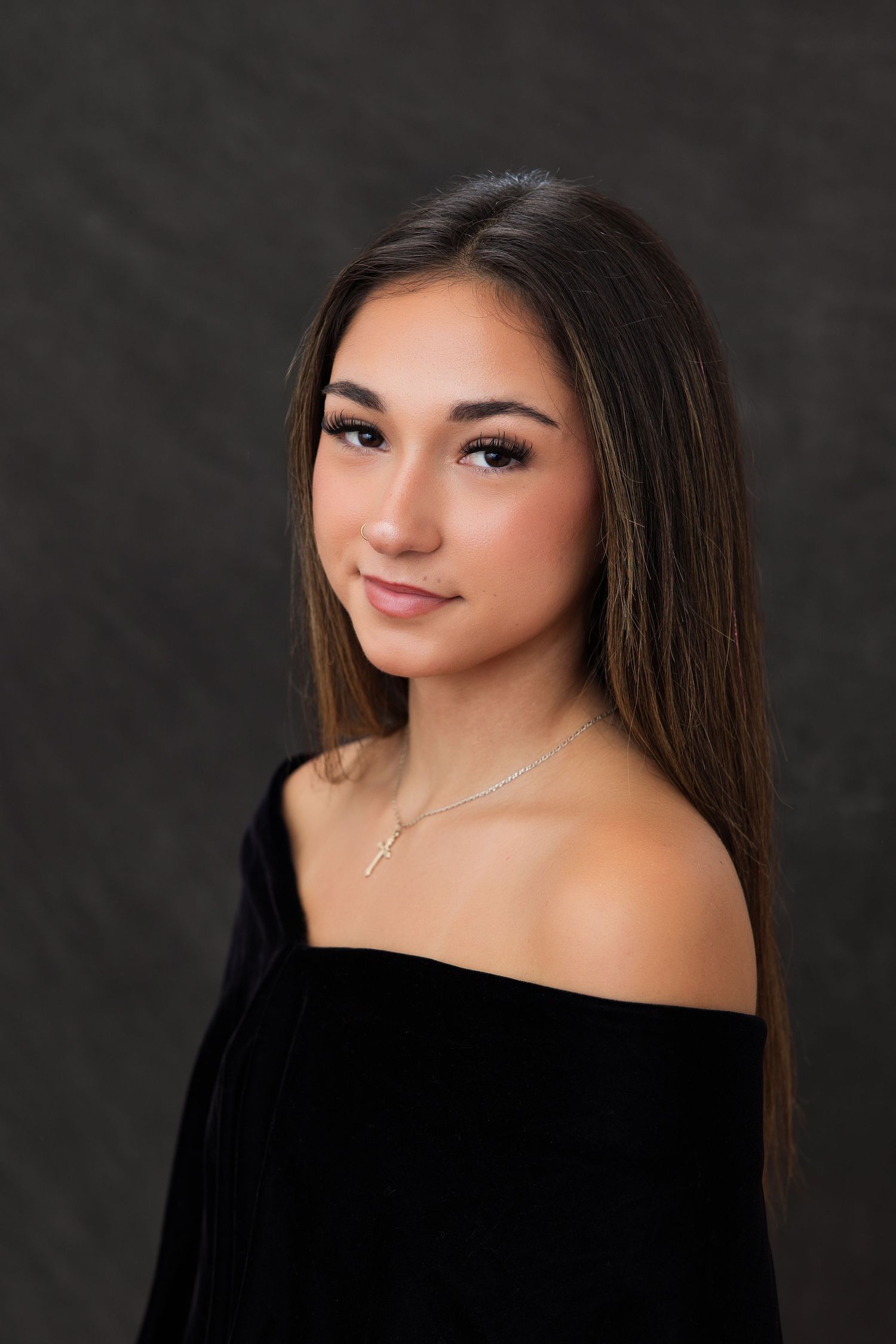 Young woman with long brown hair wearing an off-shoulder black velvet top and gold cross necklace poses for a portrait.