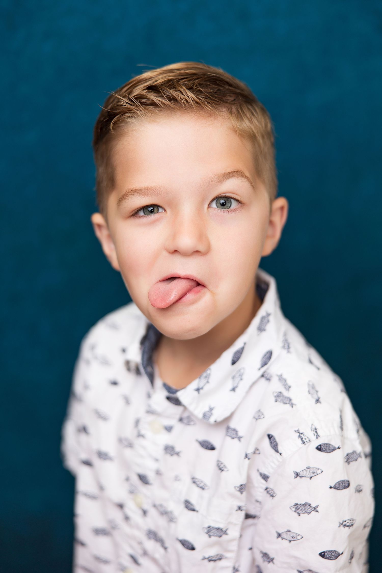 A playful young boy with blonde hair sticking out his tongue, wearing a fish-print shirt against a teal background.