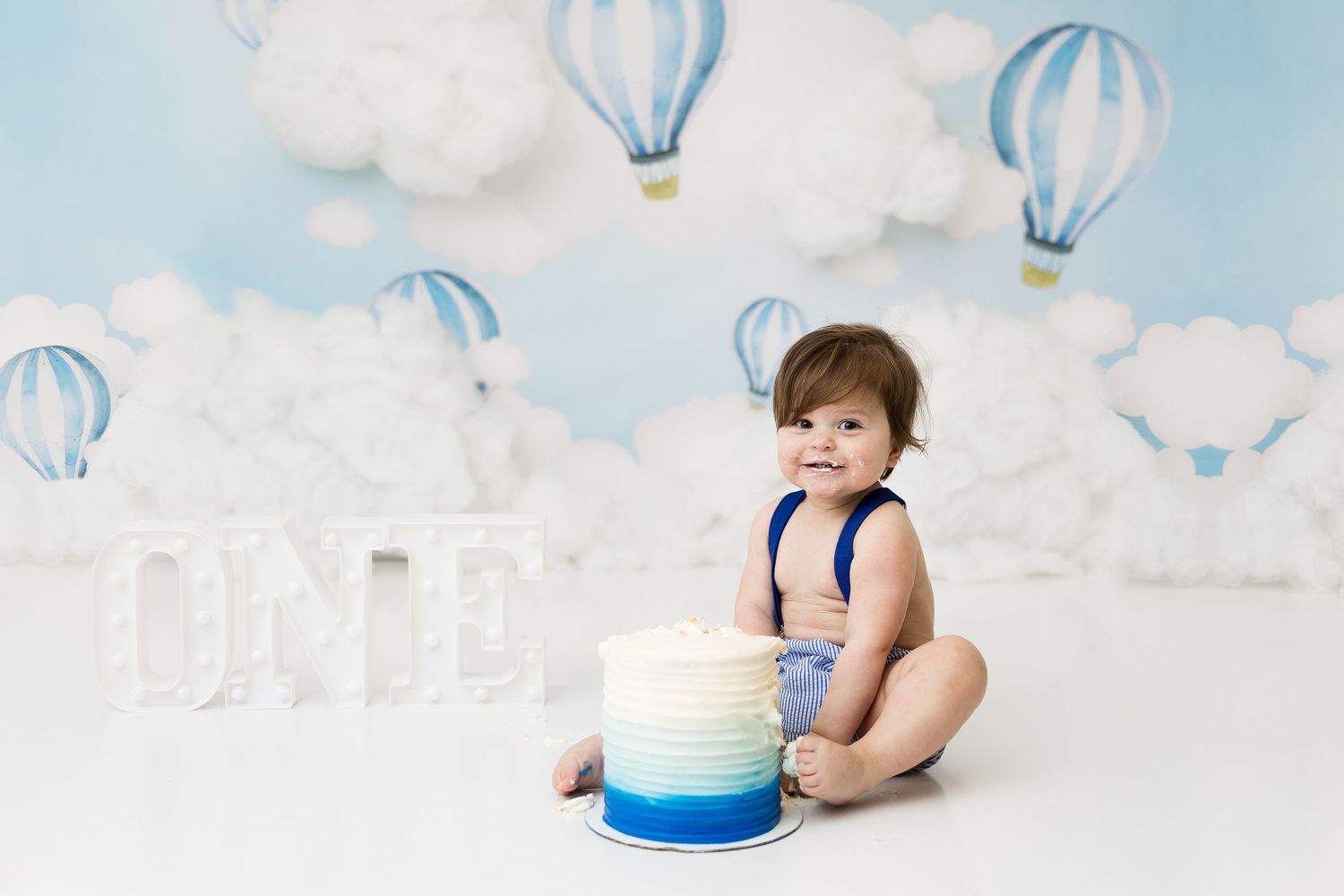 Baby in blue outfit sits with tiered cake against cloud backdrop.