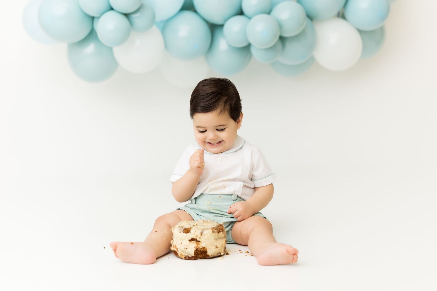 Sweet cake smash photo with baby in white and mint outfit under blue balloons.