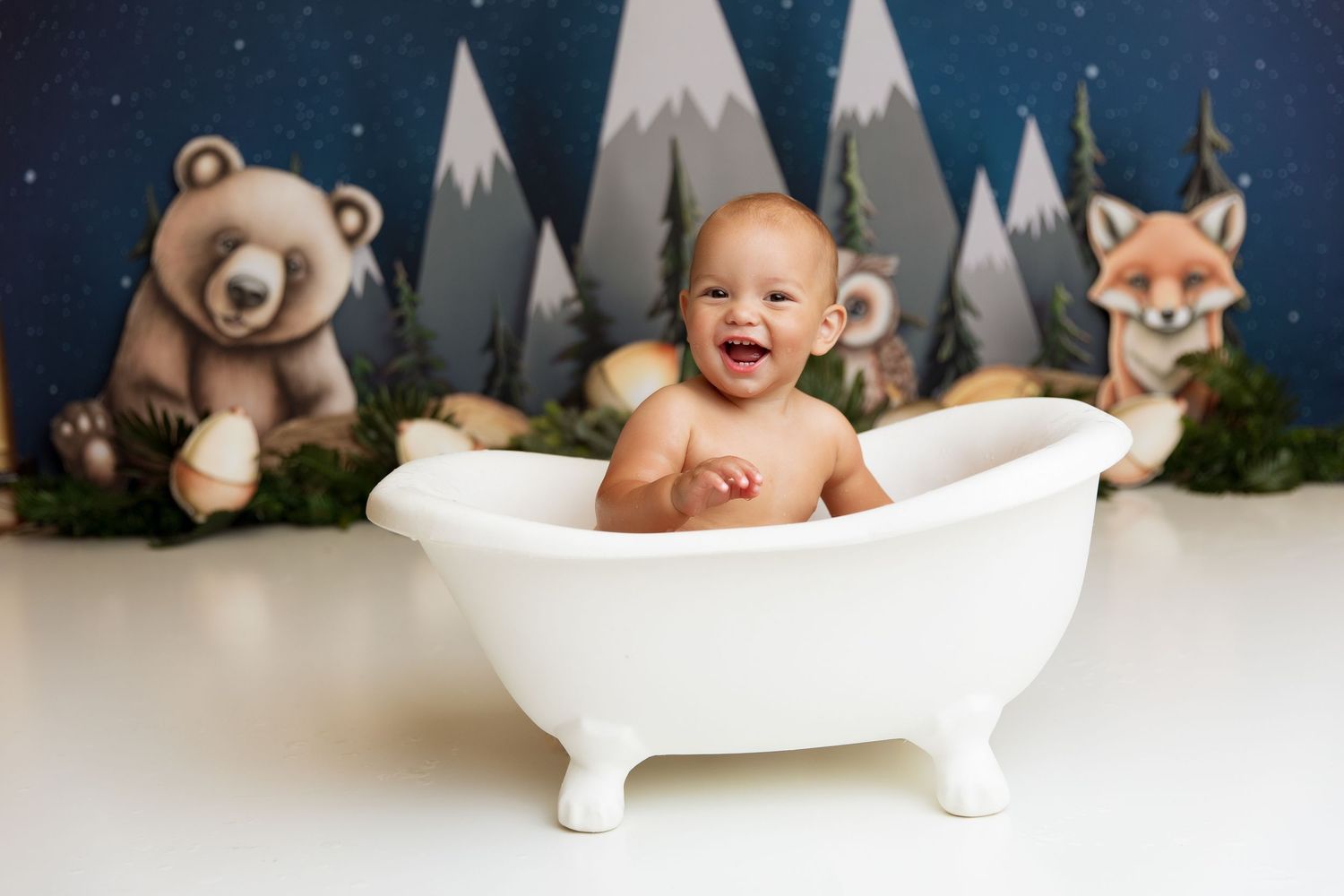Baby sits in white bathtub with woodland themed backdrop featuring bear and fox decorations.
