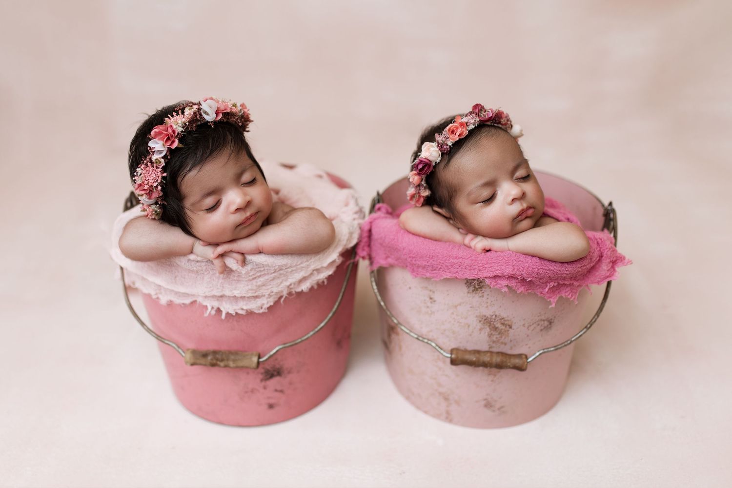 Twin newborns with floral crowns sleep in pink decorative boxes for portrait session.