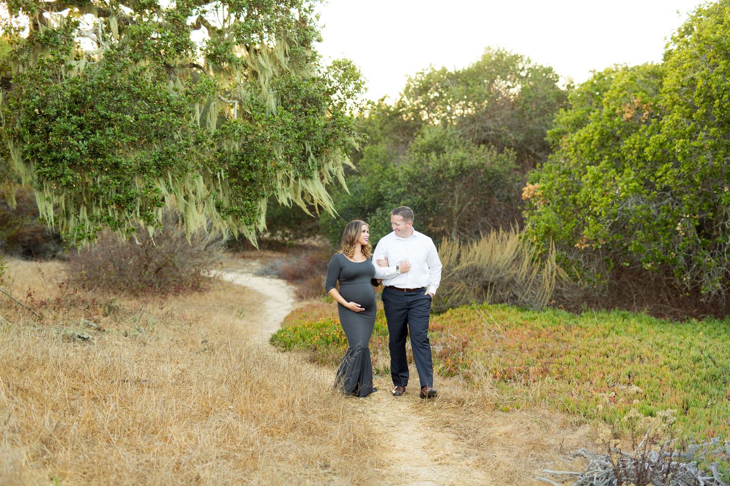 A couple strolls hand in hand along a winding dirt path through coastal vegetation.