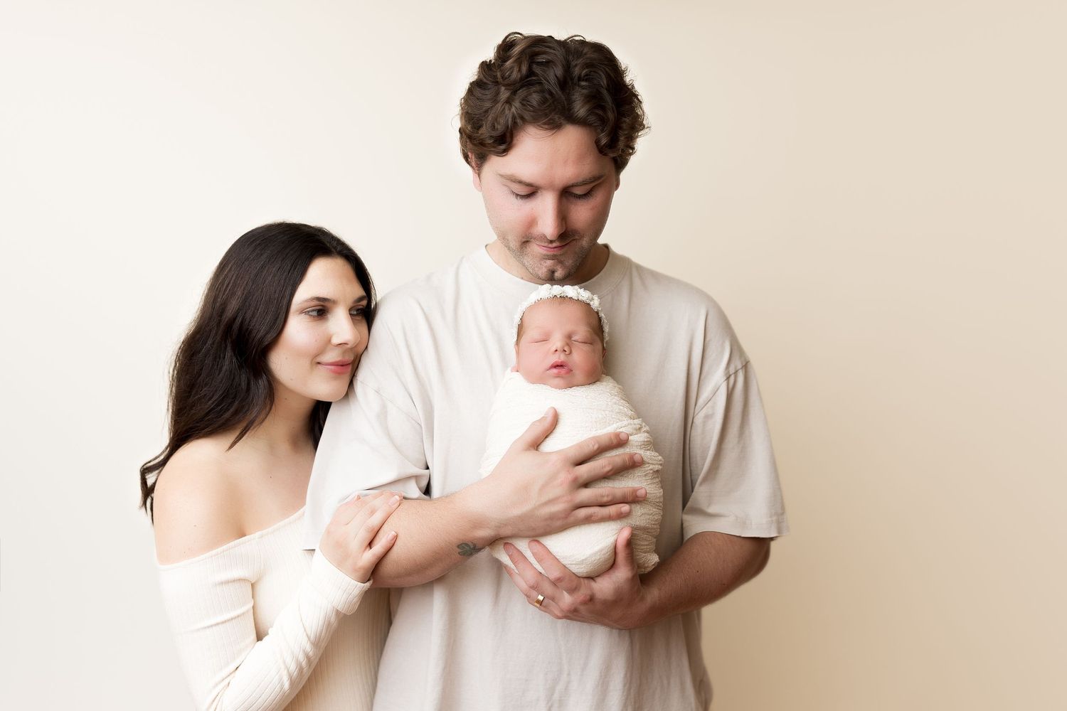 Parents in white clothing hold their sleeping newborn together against beige background.