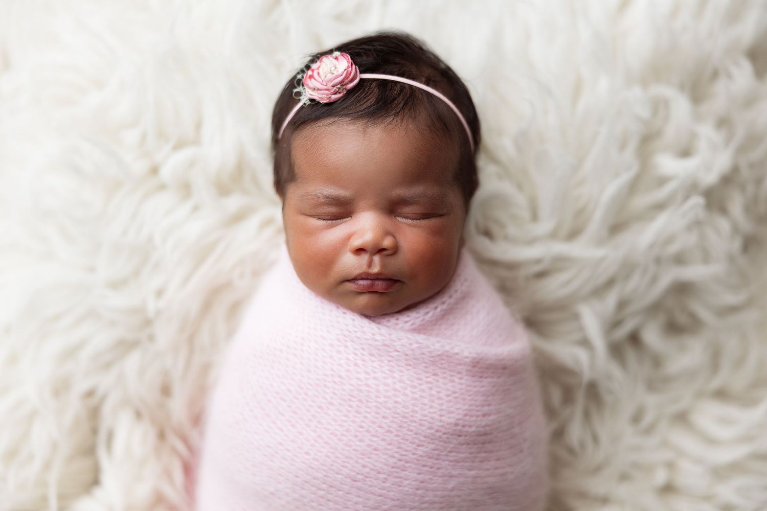 Sleeping newborn in pink wrap wearing floral headband on white fur.