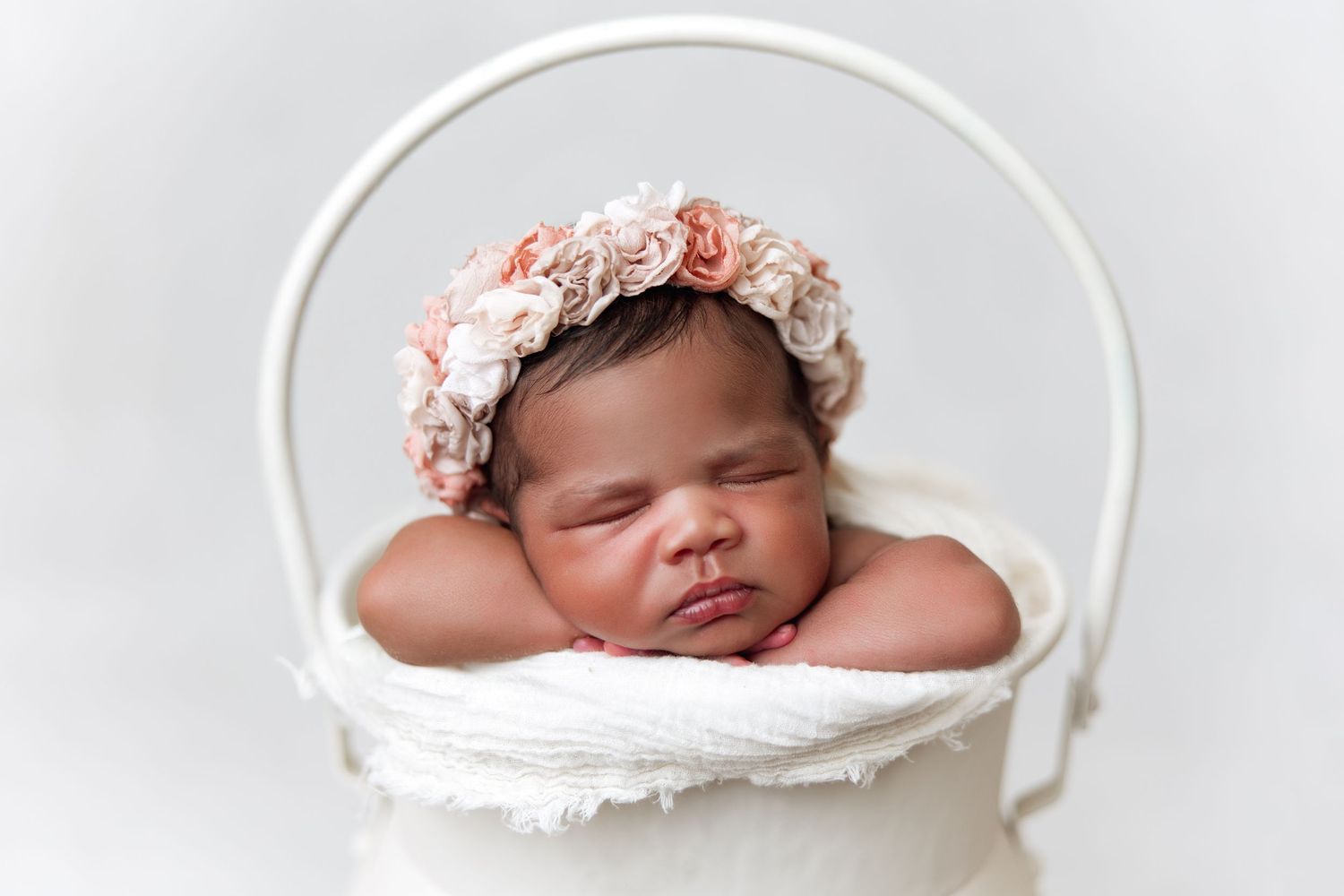 A sleeping newborn baby wearing a pink floral headband rests peacefully in a white basket against a light background.