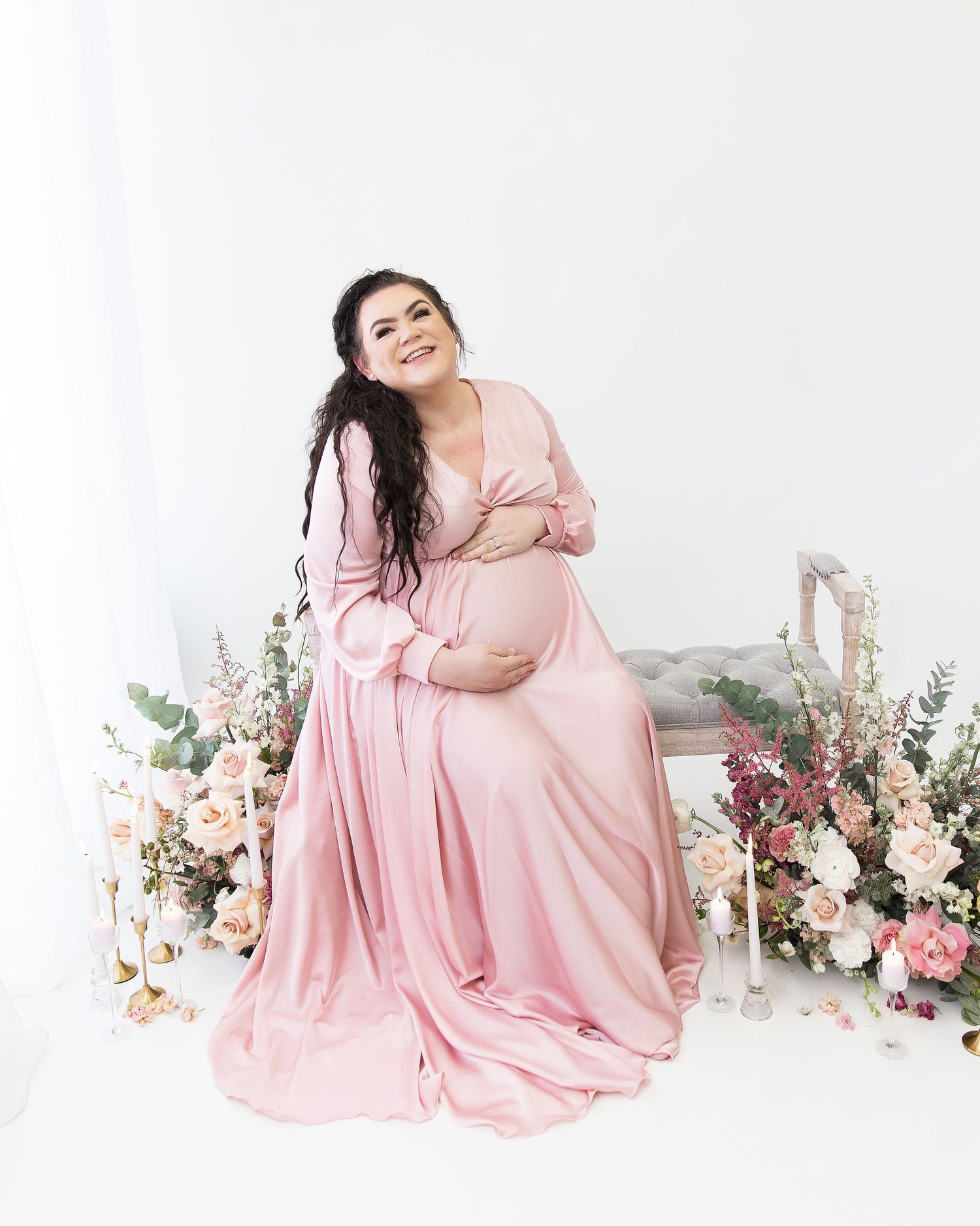 A figure in a flowing pink gown poses gracefully against a backdrop of soft pink and white floral arrangements.