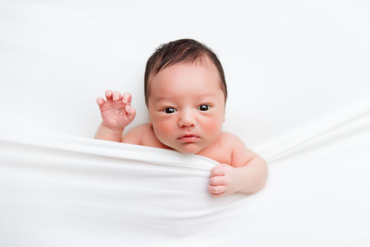 Baby peeking out from white blanket with curious expression.