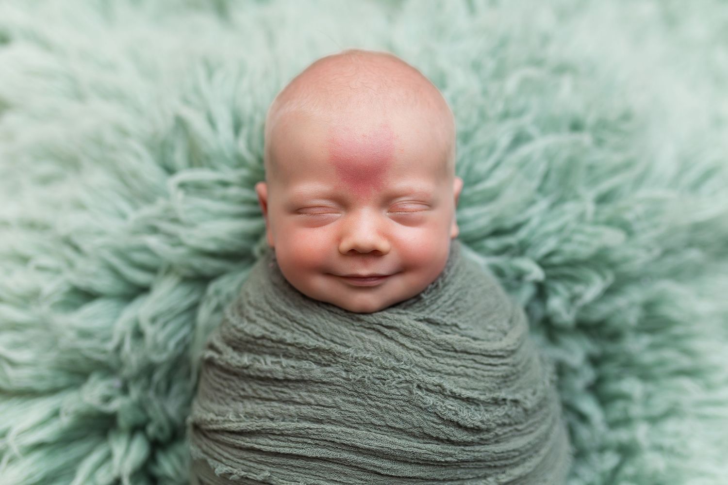 Newborn baby wrapped in sage green swaddle smiling while laying on mint colored fluffy backdrop.