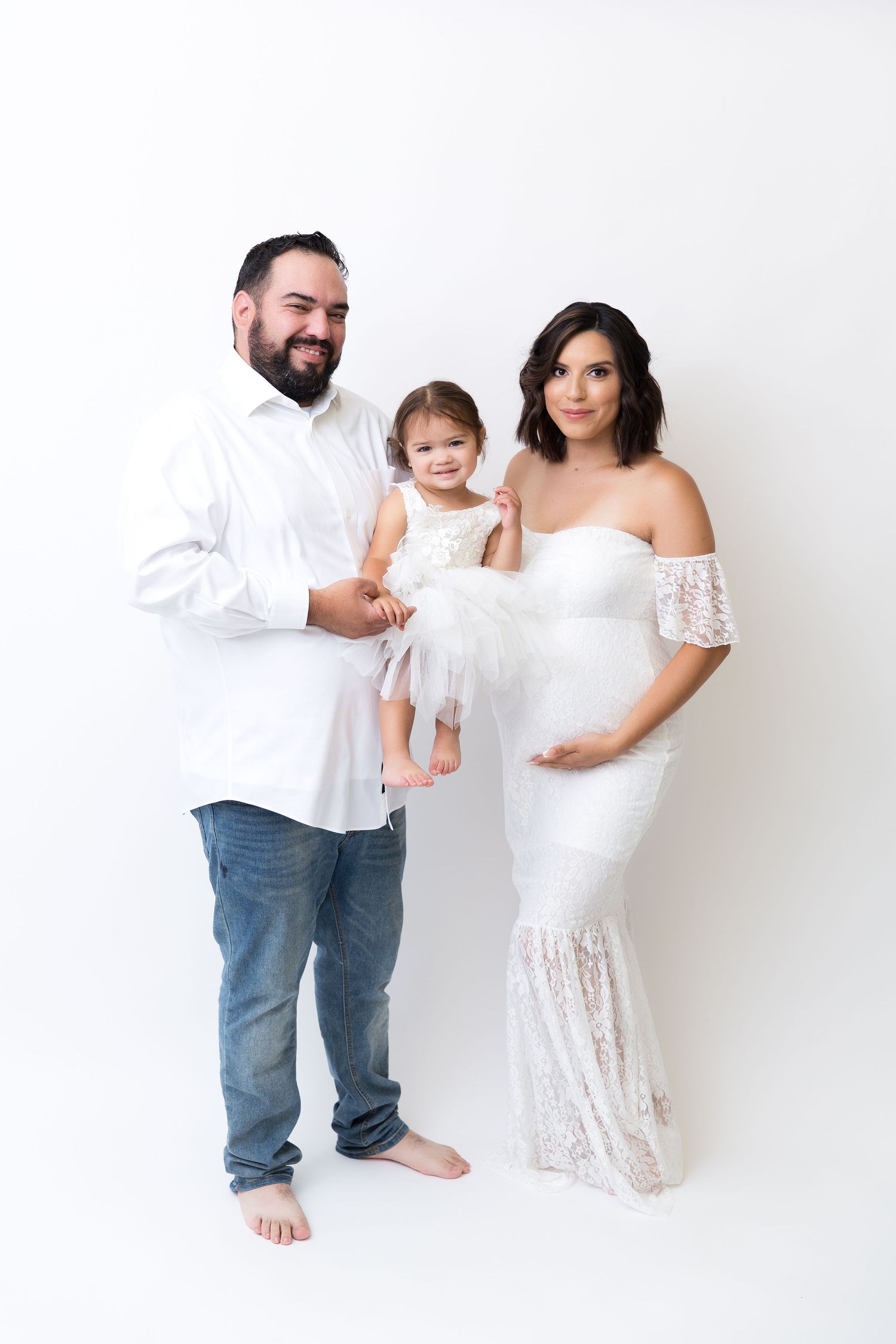 Growing family poses together in white outfits against bright backdrop.