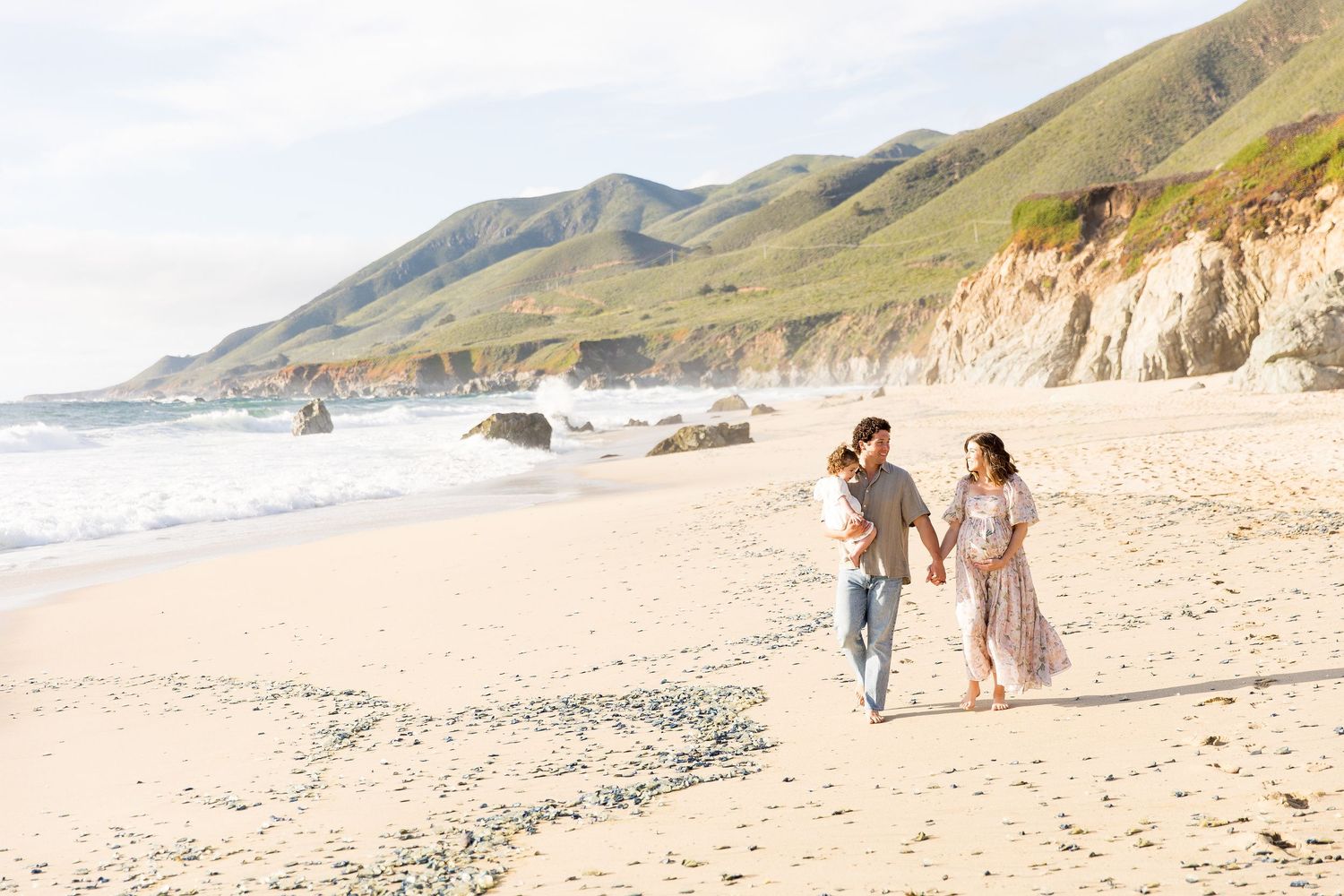Couple walks hand in hand along Garrapata beach in Big Sur coastline with mountains in background.