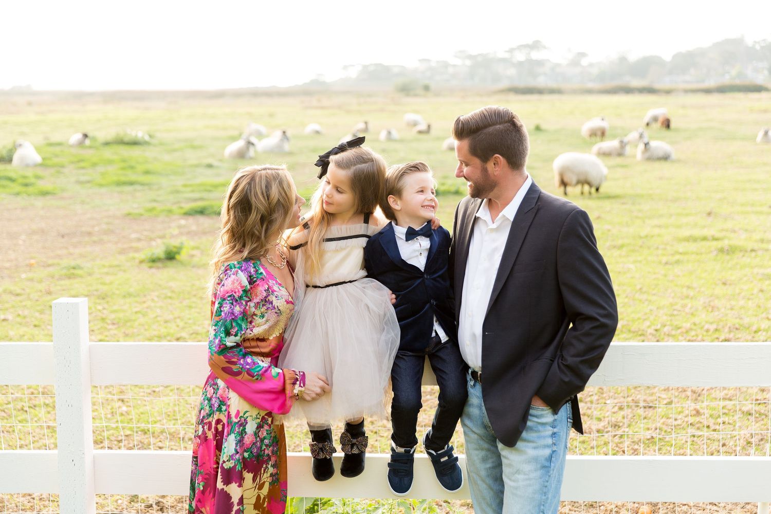 Family gathered by white fence on rural farmland with sheep grazing in background.