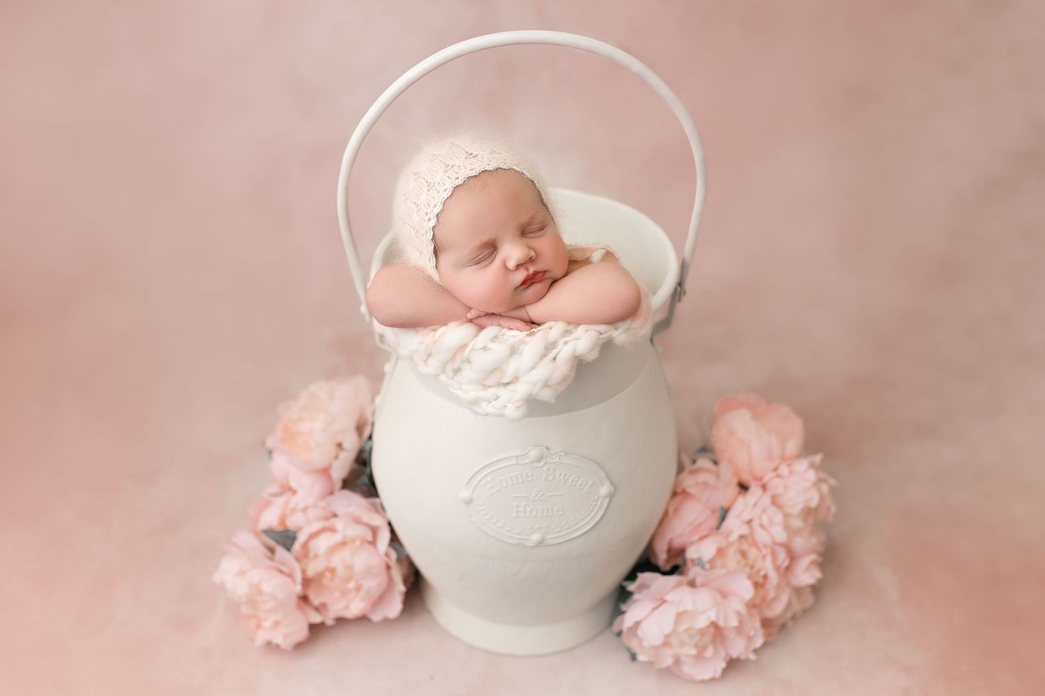 A sleeping newborn nestled in a white ceramic pot surrounded by pink flowers on a pink backdrop.