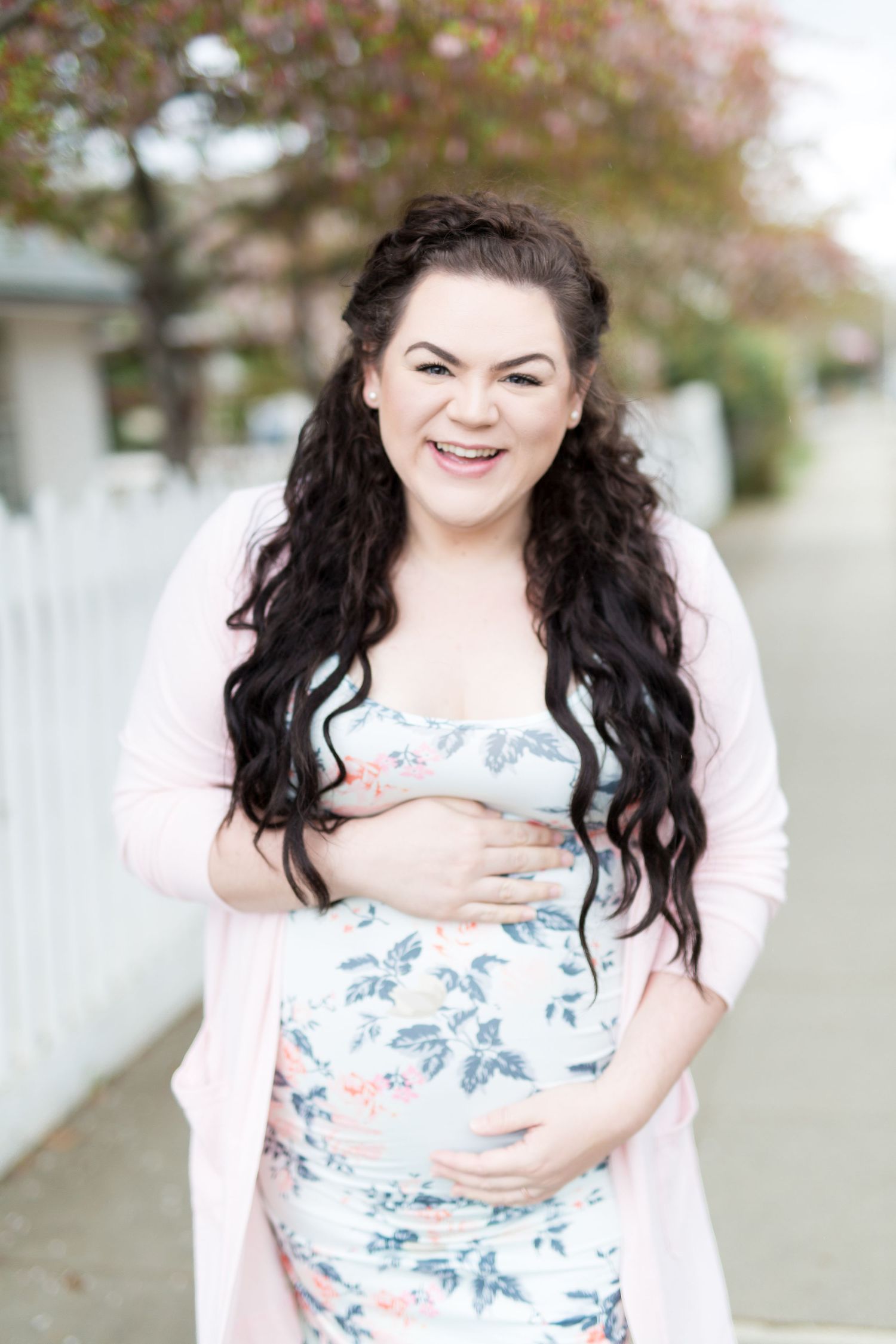 Spring maternity photos with floral dress and pink cardigan against white fence.