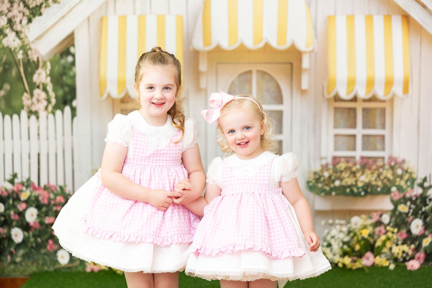 Two adorable pink dresses shown in front of a yellow and white playhouse with garden backdrop.