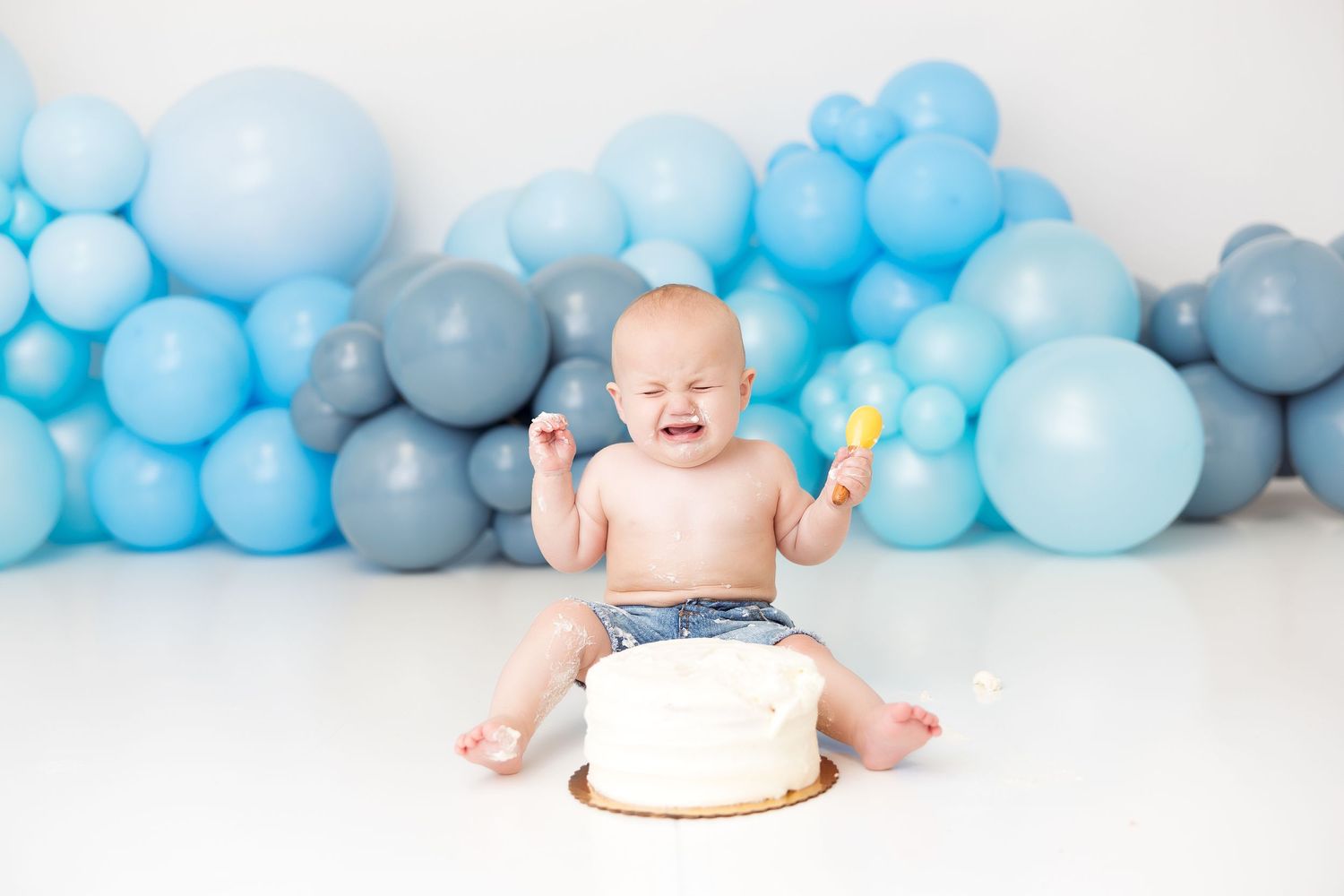 A baby enjoying a smash cake celebration against a backdrop of blue and gray balloons.