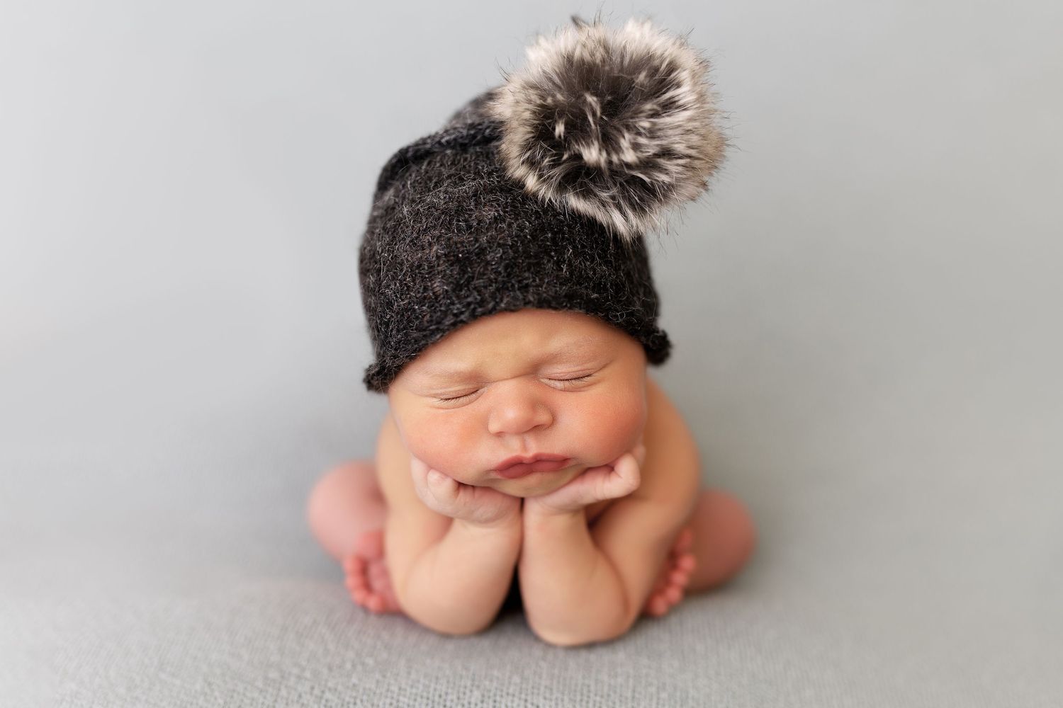 A sleeping newborn posed with hands under chin wearing a dark knit hat with fur pom pom.
