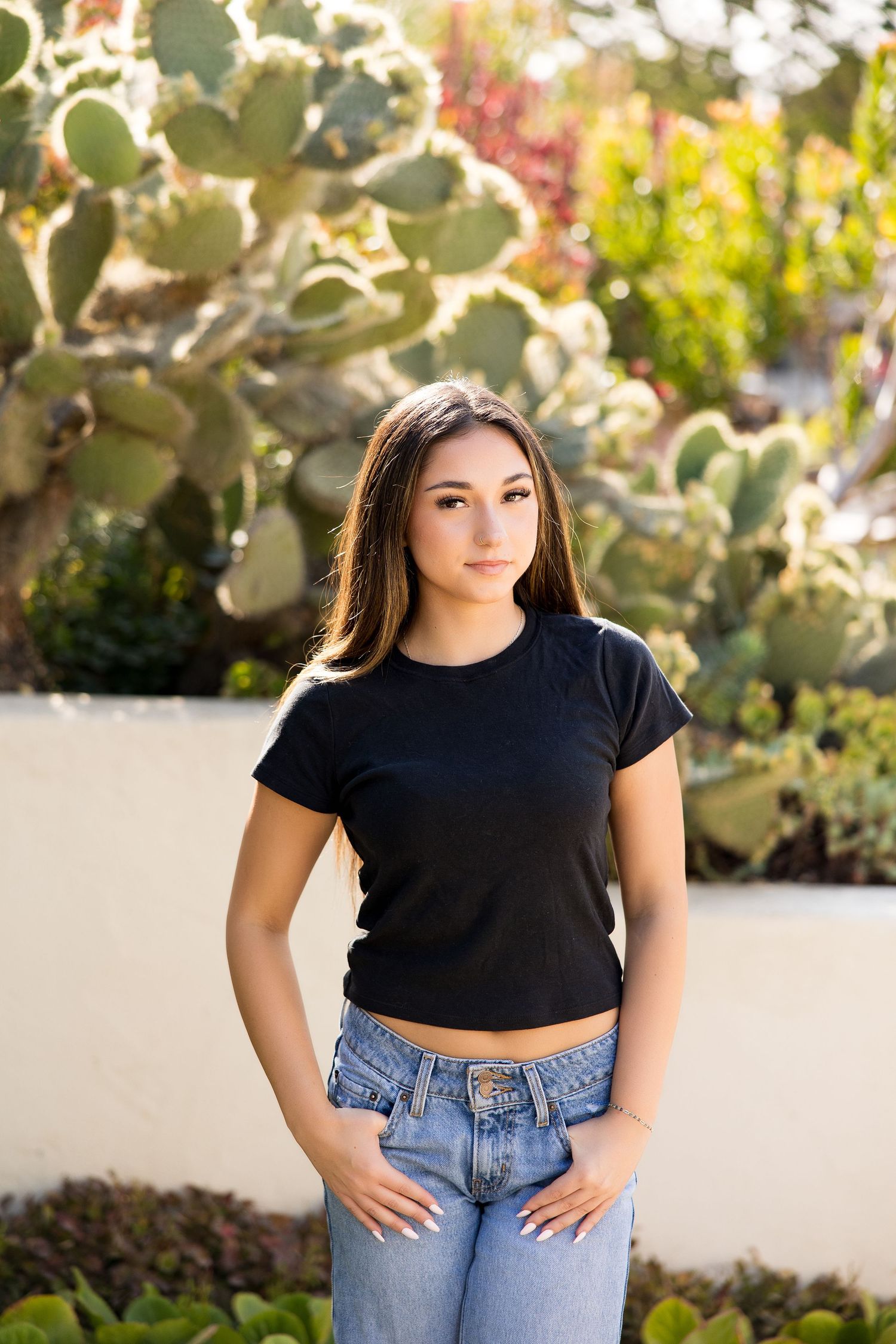 Young woman in black crop top and jeans posing outdoors in front of large prickly pear cactus garden.