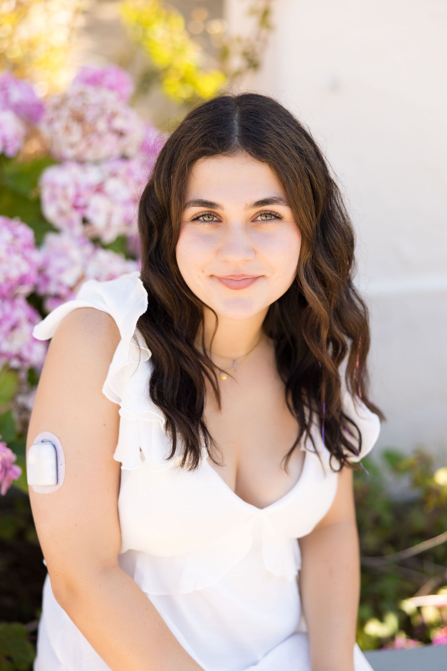 Young woman in white dress smiling outdoors with pink flowers, showing a medical patch on her upper arm.