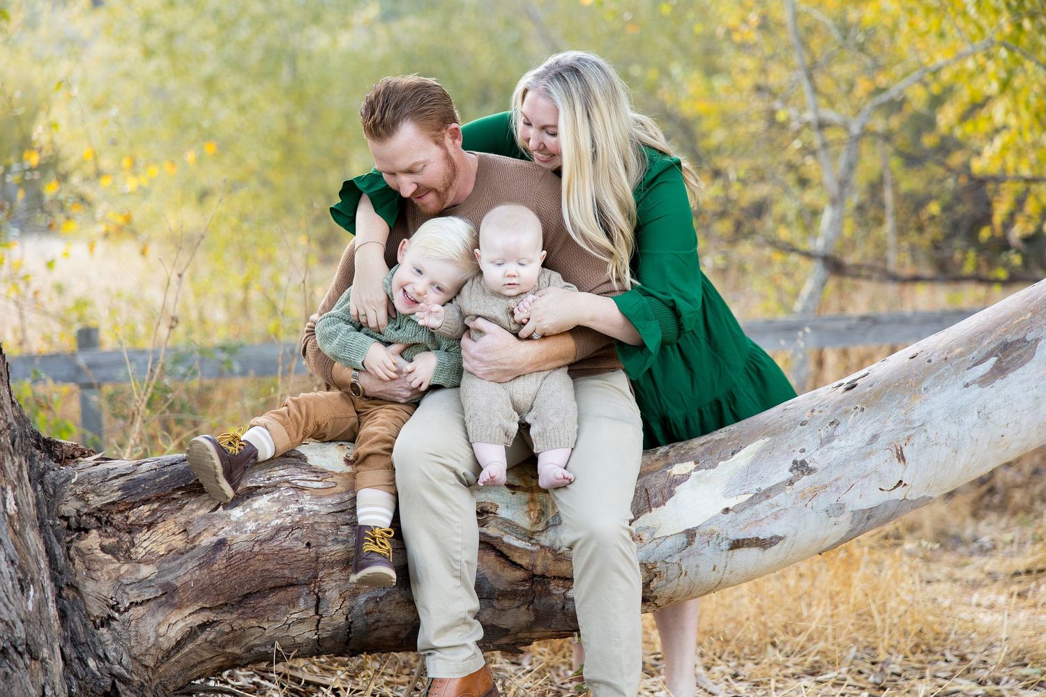 Family photography session on fallen tree trunk with autumn foliage in background.