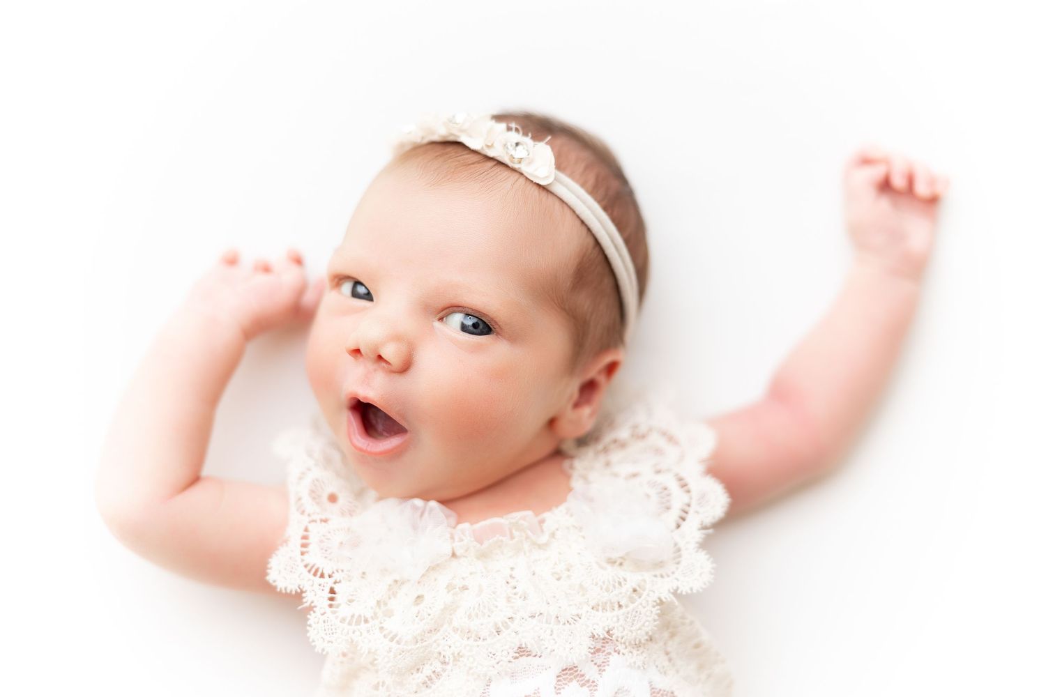 Alert baby in lace dress and headband stretches arms against white background.