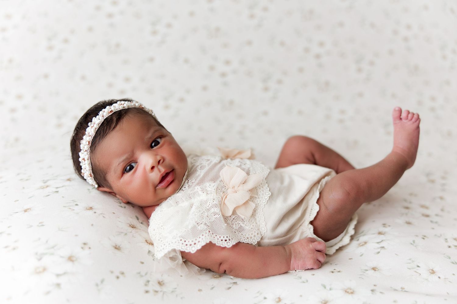 Infant wearing white lace dress and headband lying on polka dot backdrop.