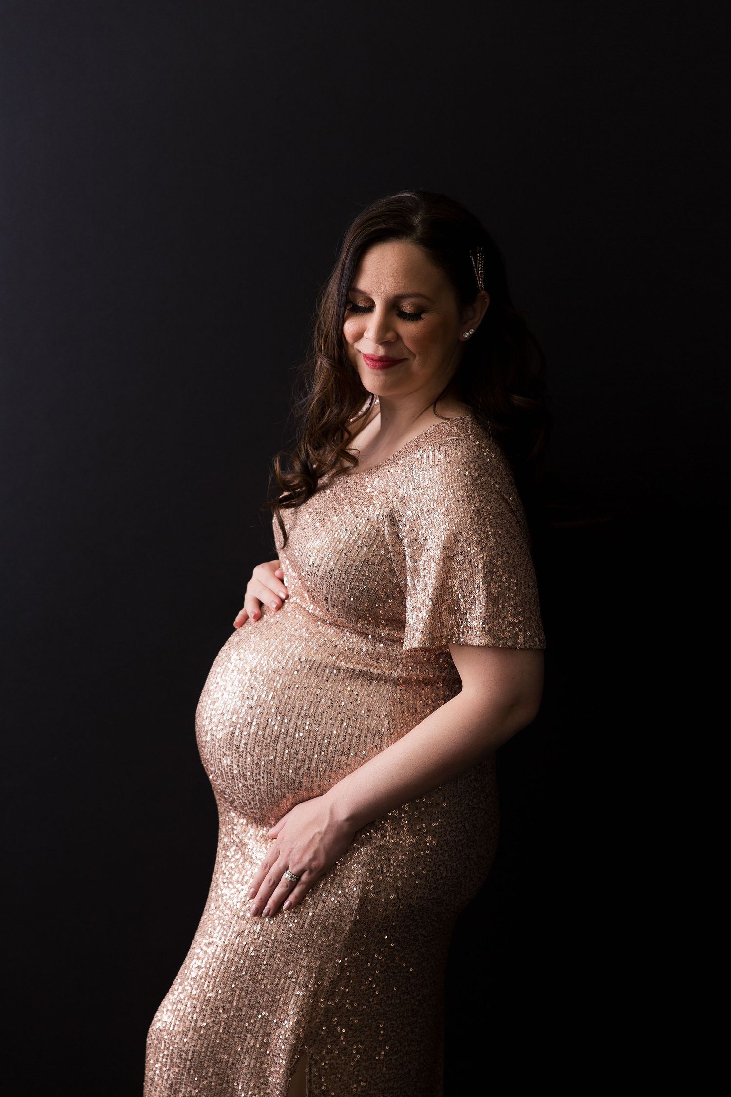 Elegant maternity portrait featuring sparkly rose gold dress against dark background.