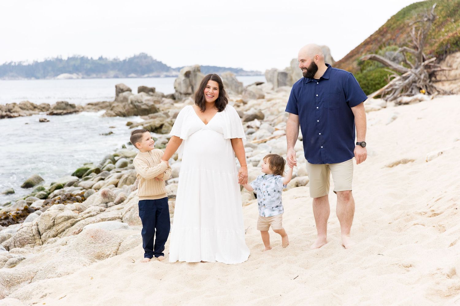 Family walks together along scenic beach with rocky coastline and mountains in background.