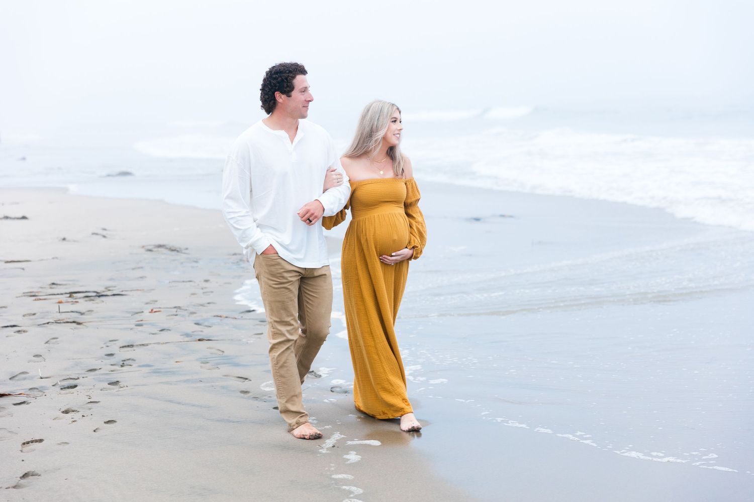 Expecting couple walking barefoot along ocean shoreline during cloudy beach session.