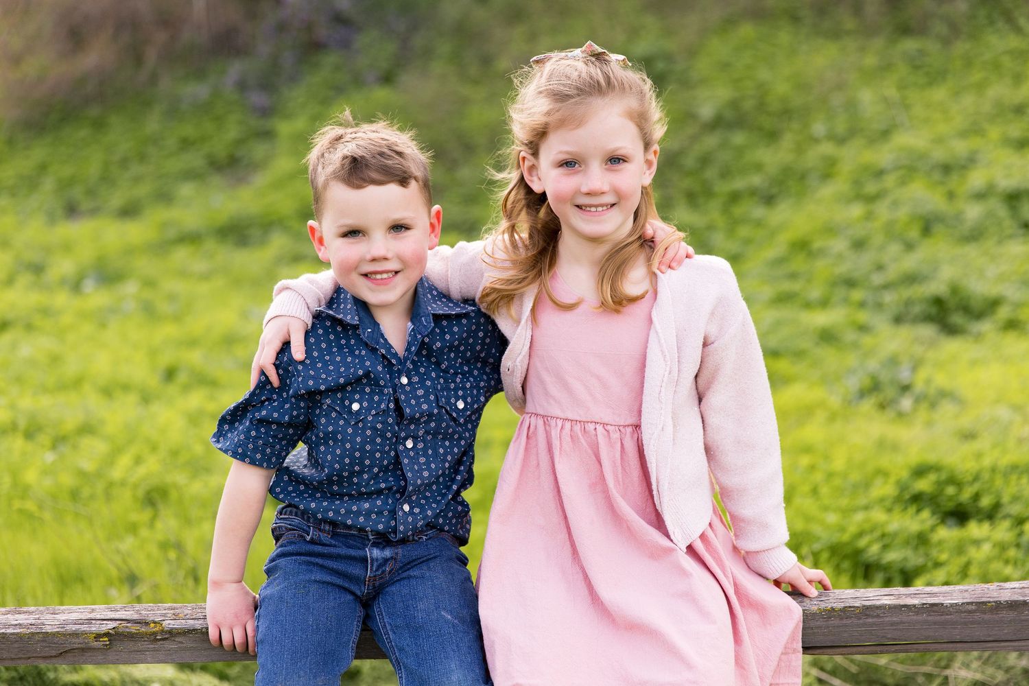 Brother and sister smiling together by a wooden fence in green hills during a Hollister family photo session.