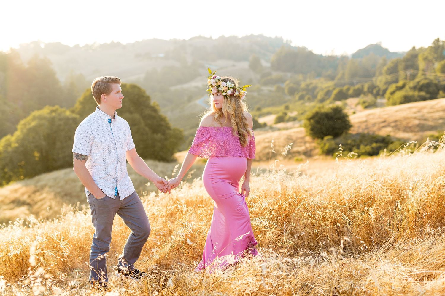 A maternity couple holds hands while walking through a golden field at sunset in the rolling hills of San Jose.