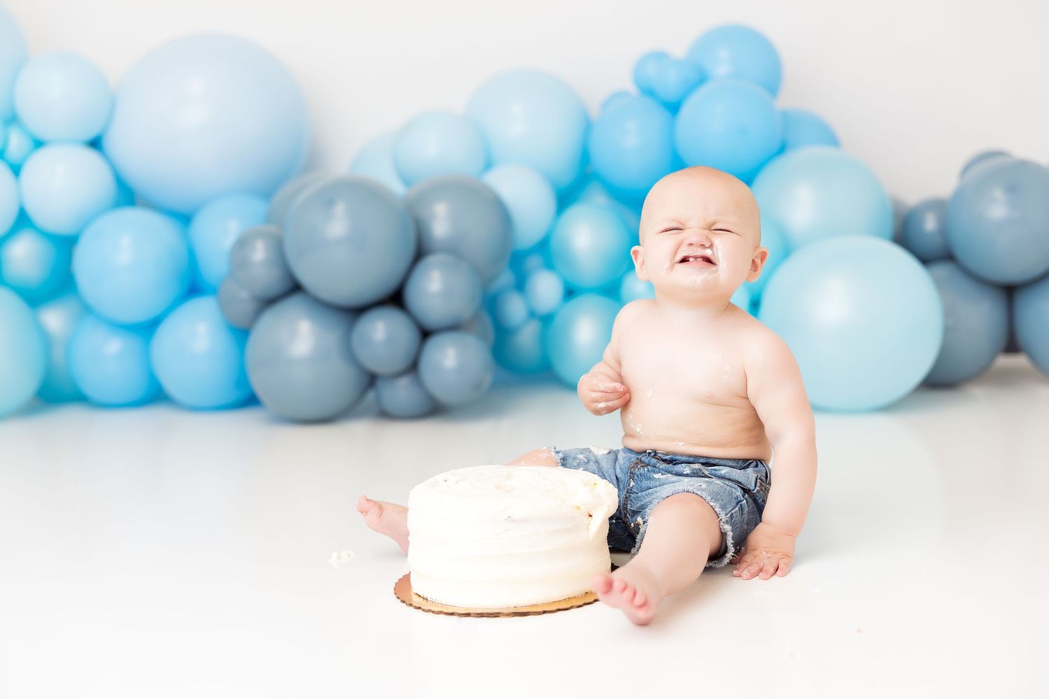 Baby having fun with cake smash photography session surrounded by blue balloons and decorations.