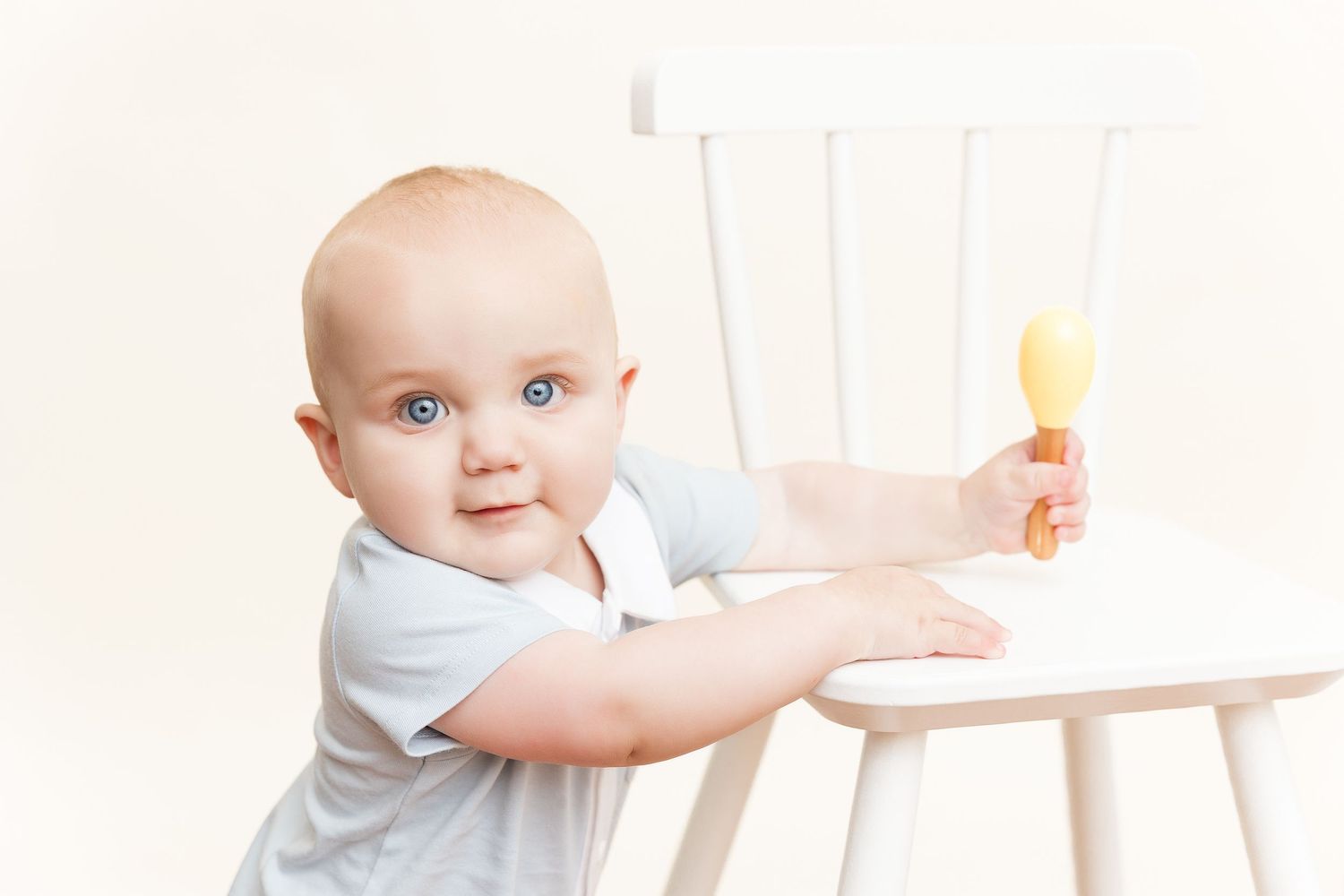 A small child in a white shirt holds onto a white chair while looking directly at the camera.