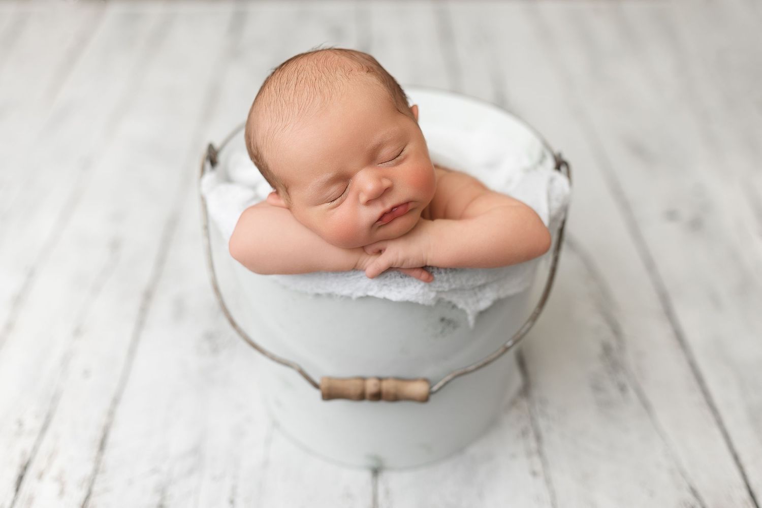 A sleeping newborn baby swaddled in white fabric rests peacefully in a metal bowl against a white backdrop.