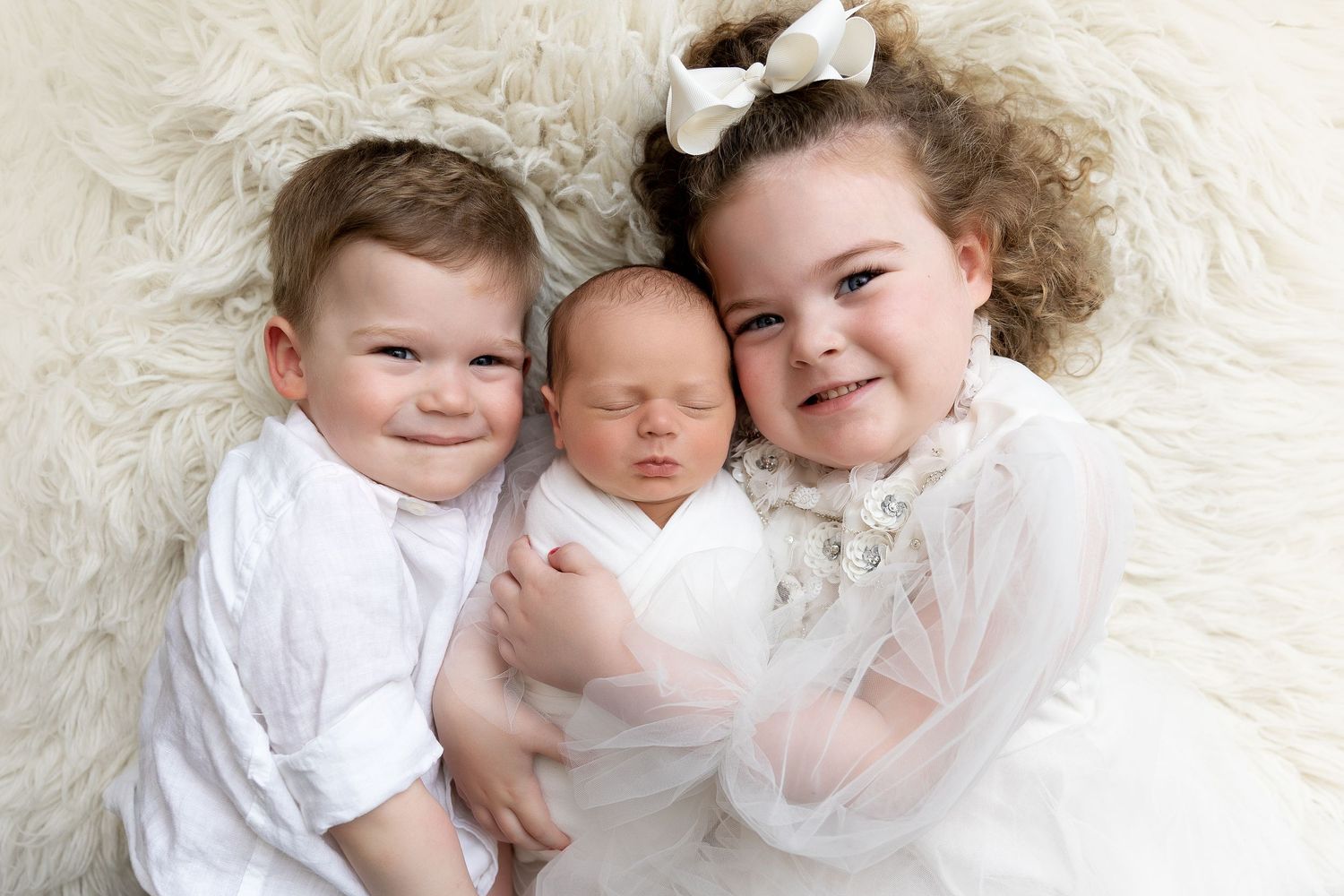 Three young children in white clothing lay together on a soft white textured surface.