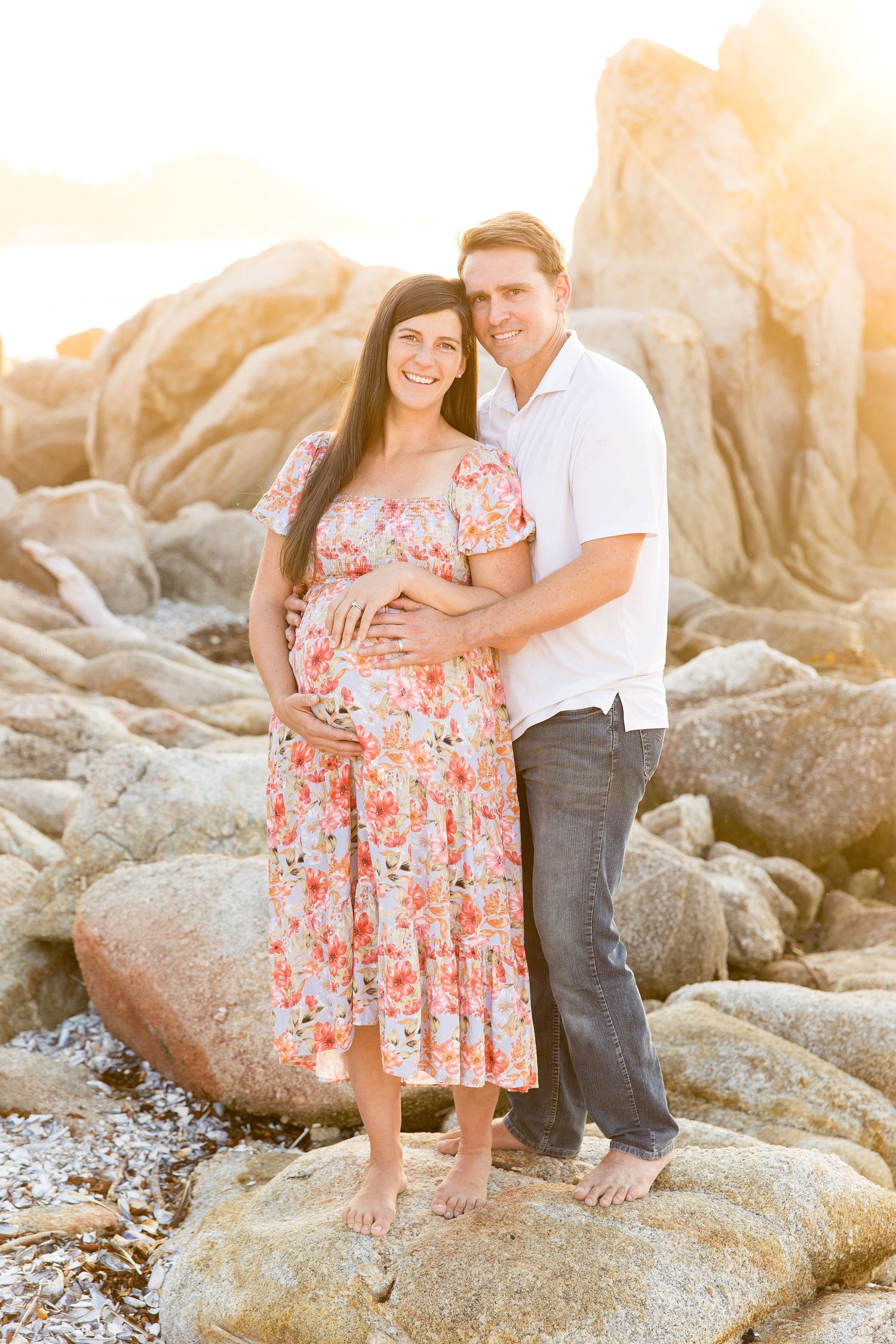 Expectant couple posing on rocky beach during sunset photoshoot.