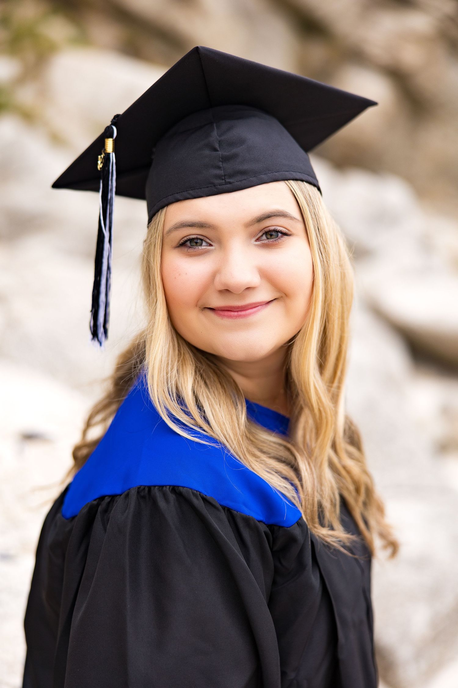 Young blonde woman smiling in black graduation cap and gown with blue trim, posing outdoors near rocks.