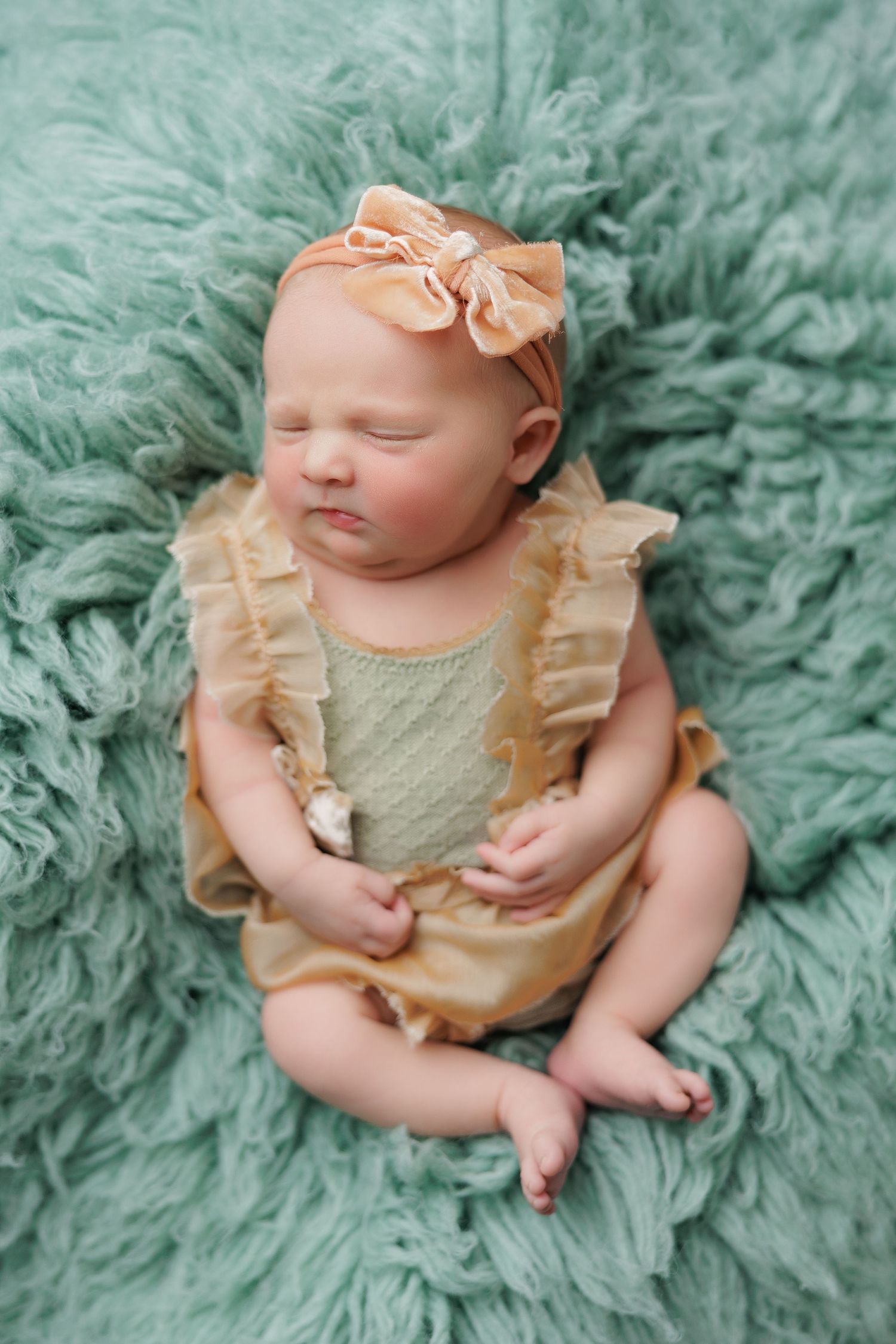 Sleeping newborn baby girl in golden ruffle romper and bow headband resting on soft teal flur rug.