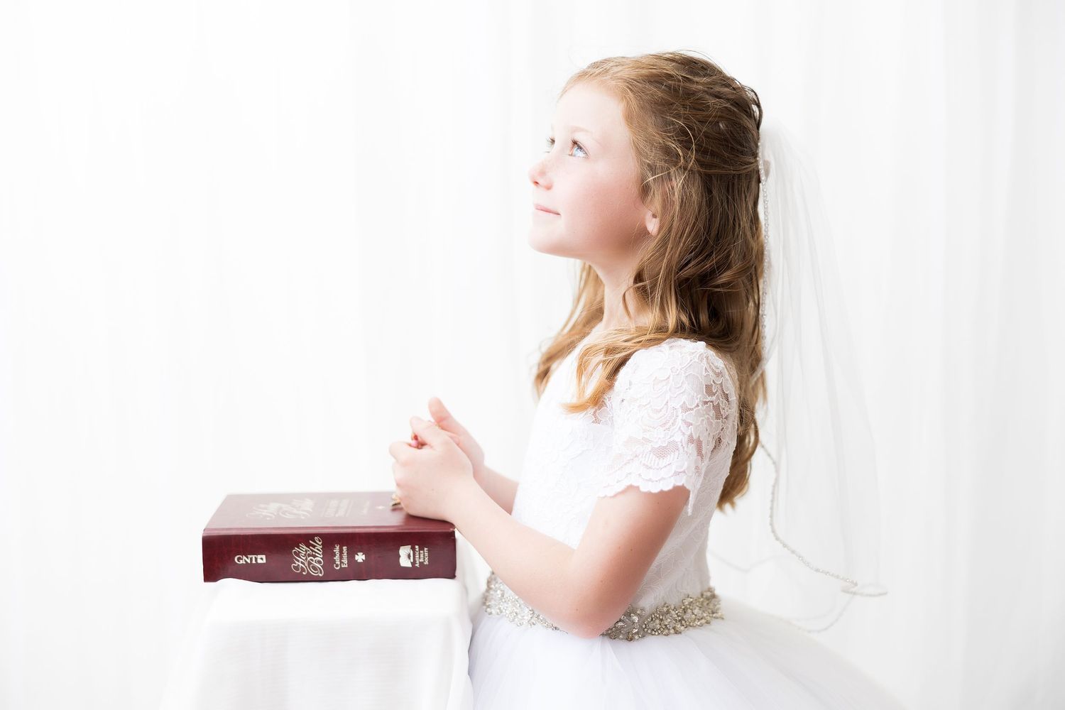 A child in first communion attire looking upward while resting hands on a Bible.