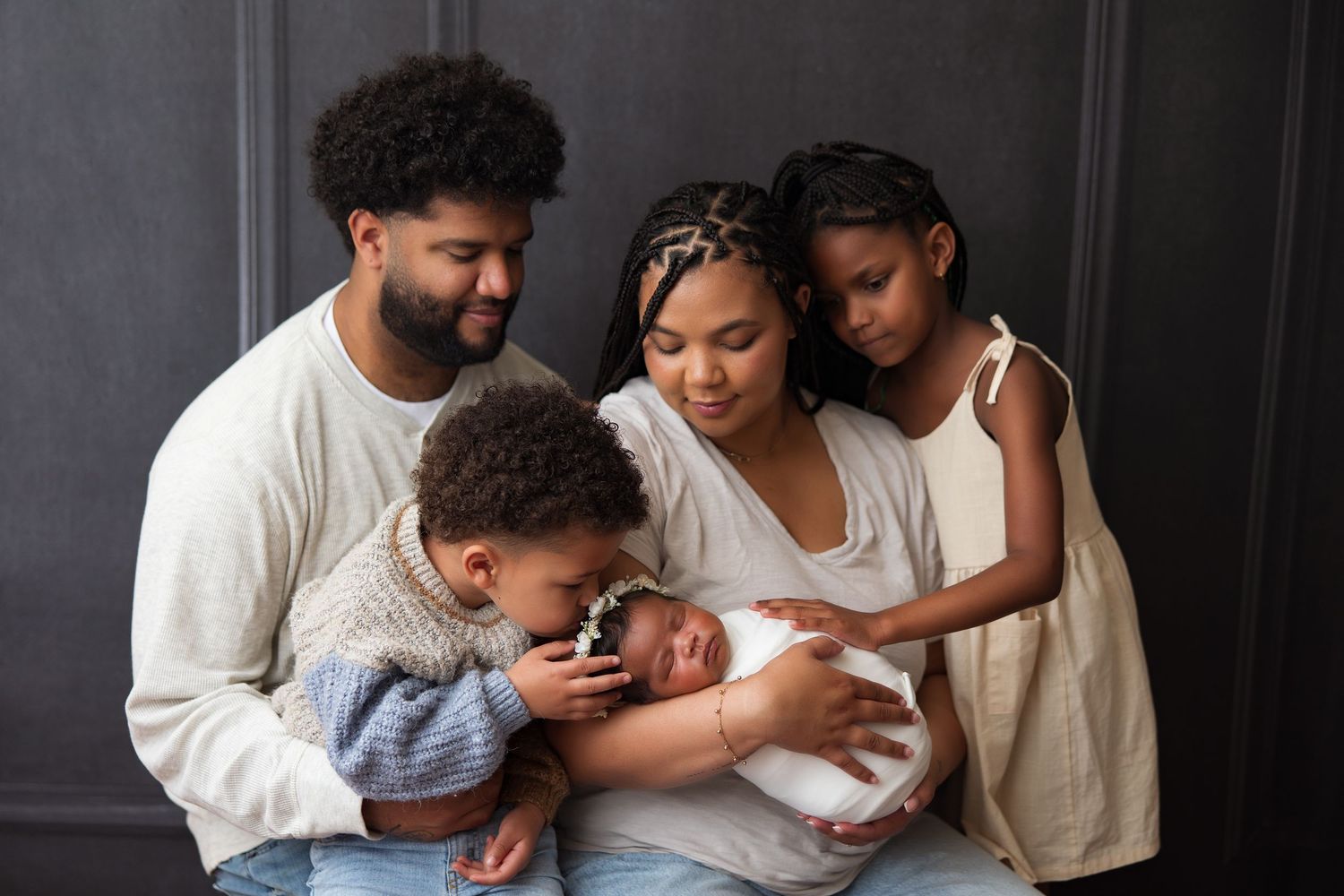 Family gathering around newborn baby against dark grey wall.