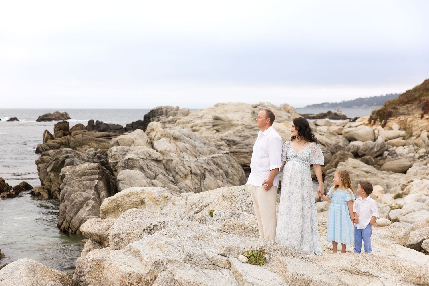 Family stands together on dramatic rocky coastal outcrop during sunset photoshoot.