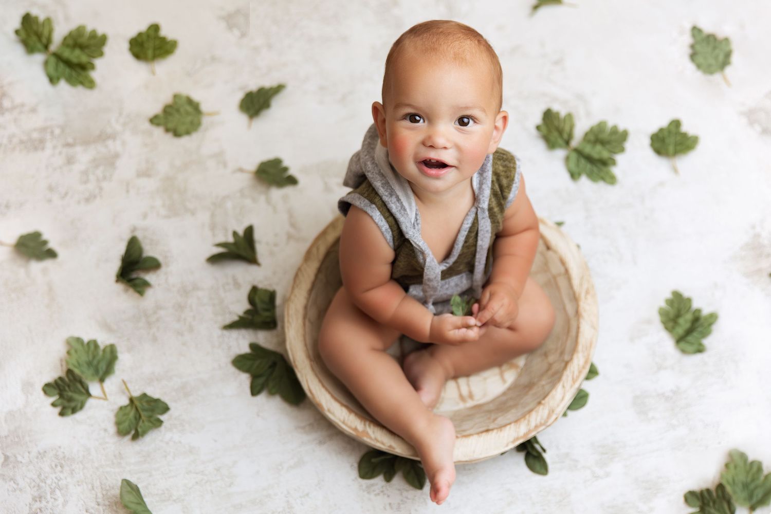 Baby wearing grey overalls sits in wooden bowl surrounded by green leaf decorations on white background.