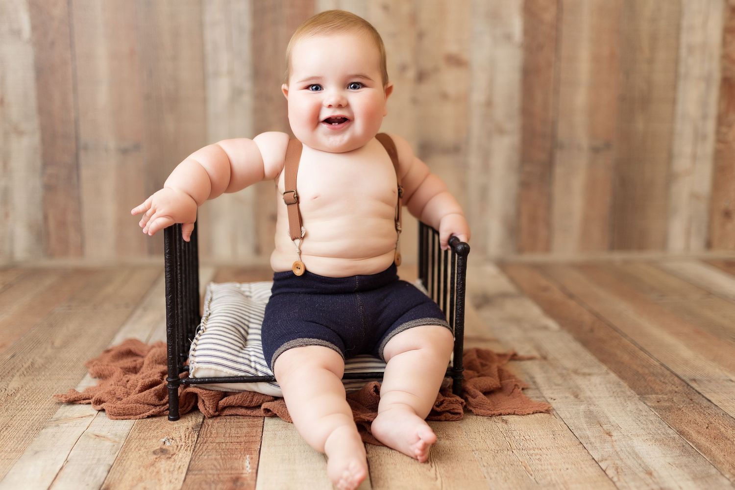 Happy baby in navy shorts and suspenders sits on dark wooden chair against wooden wall.