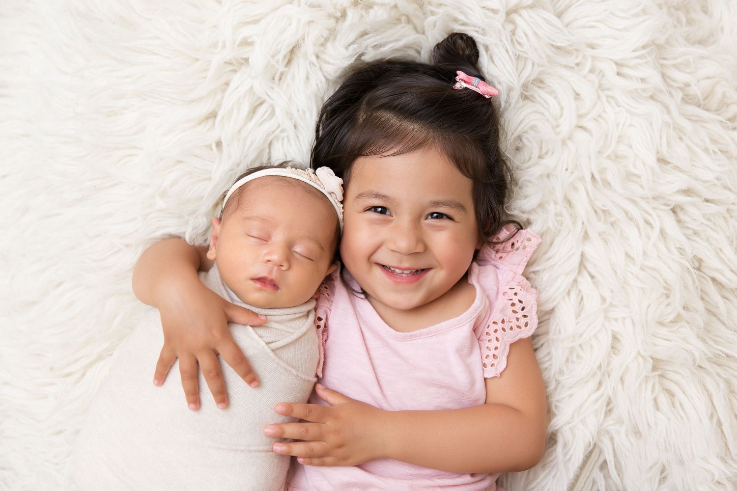 Joyful sibling portrait on fluffy white rug with sleeping newborn and smiling sister.