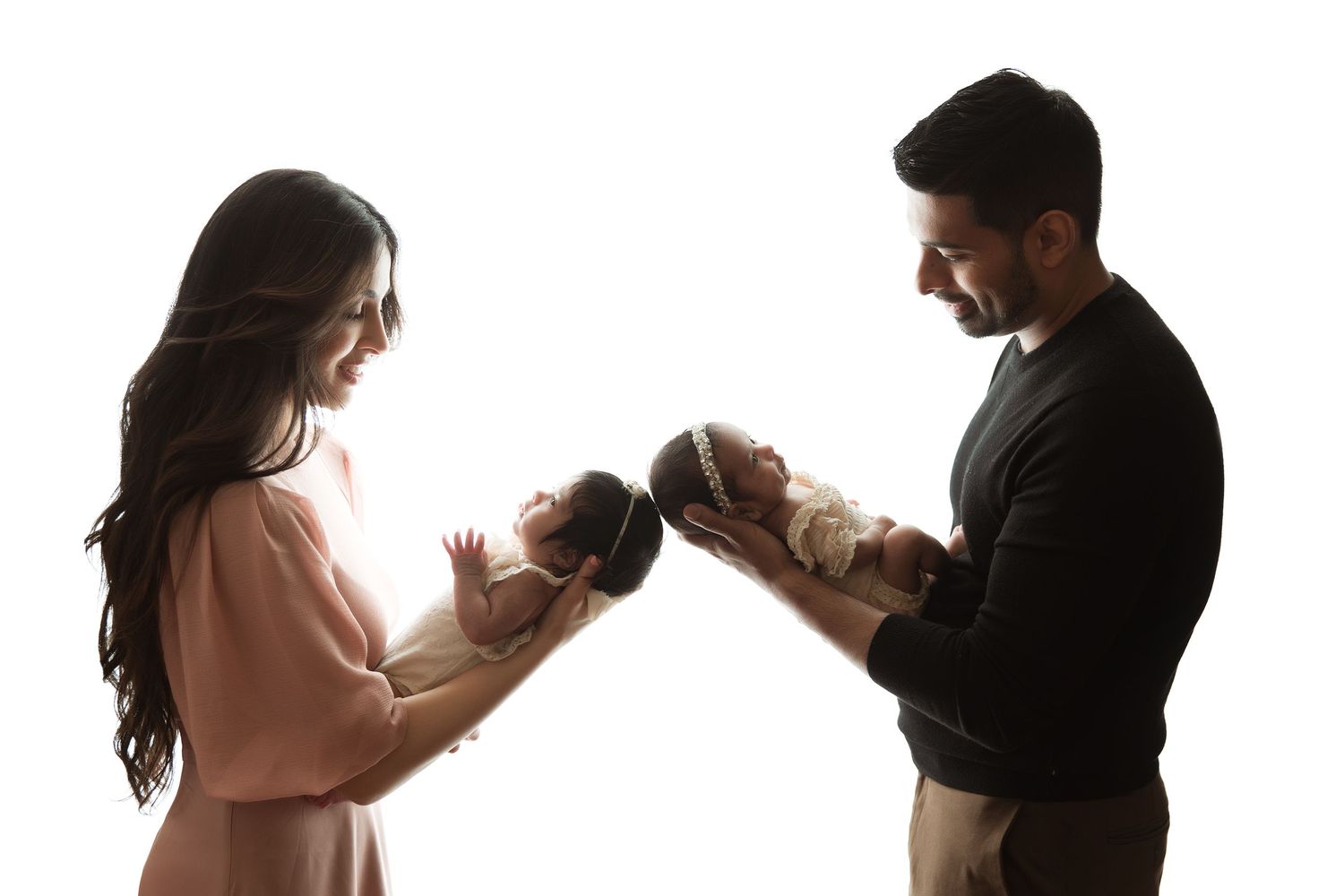 Parents in silhouette hold and gaze lovingly at their twin babies against white background.