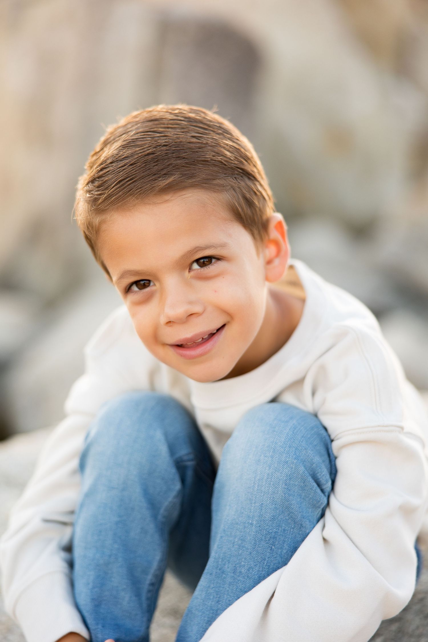 A child wearing a white sweater and jeans sitting casually on rocks in natural lighting.