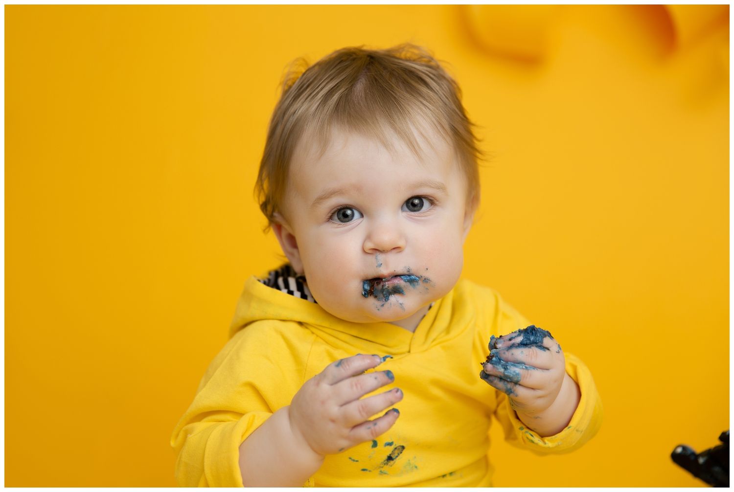 Baby wearing yellow shirt holds and eats blue cookie against vibrant yellow background.