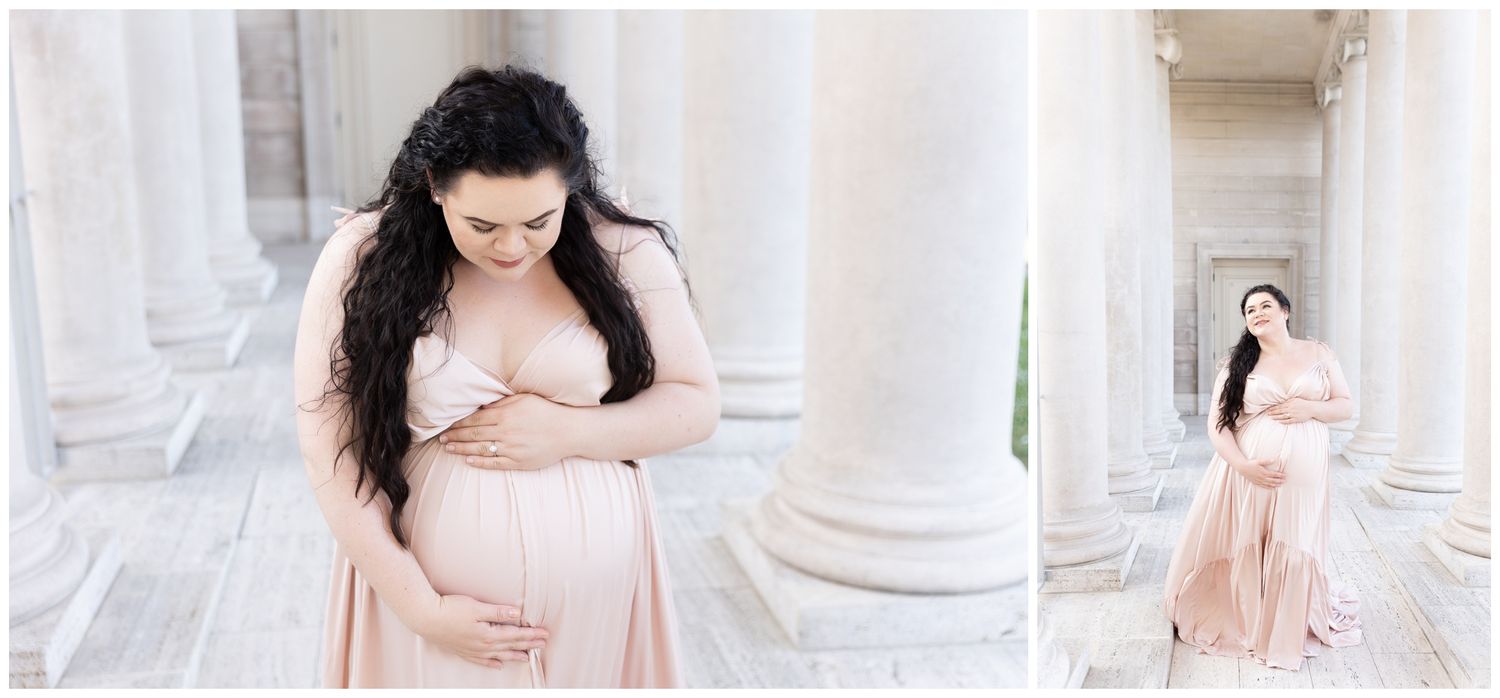 A woman in a flowing pink dress poses elegantly against white stone columns in a maternity photo session.