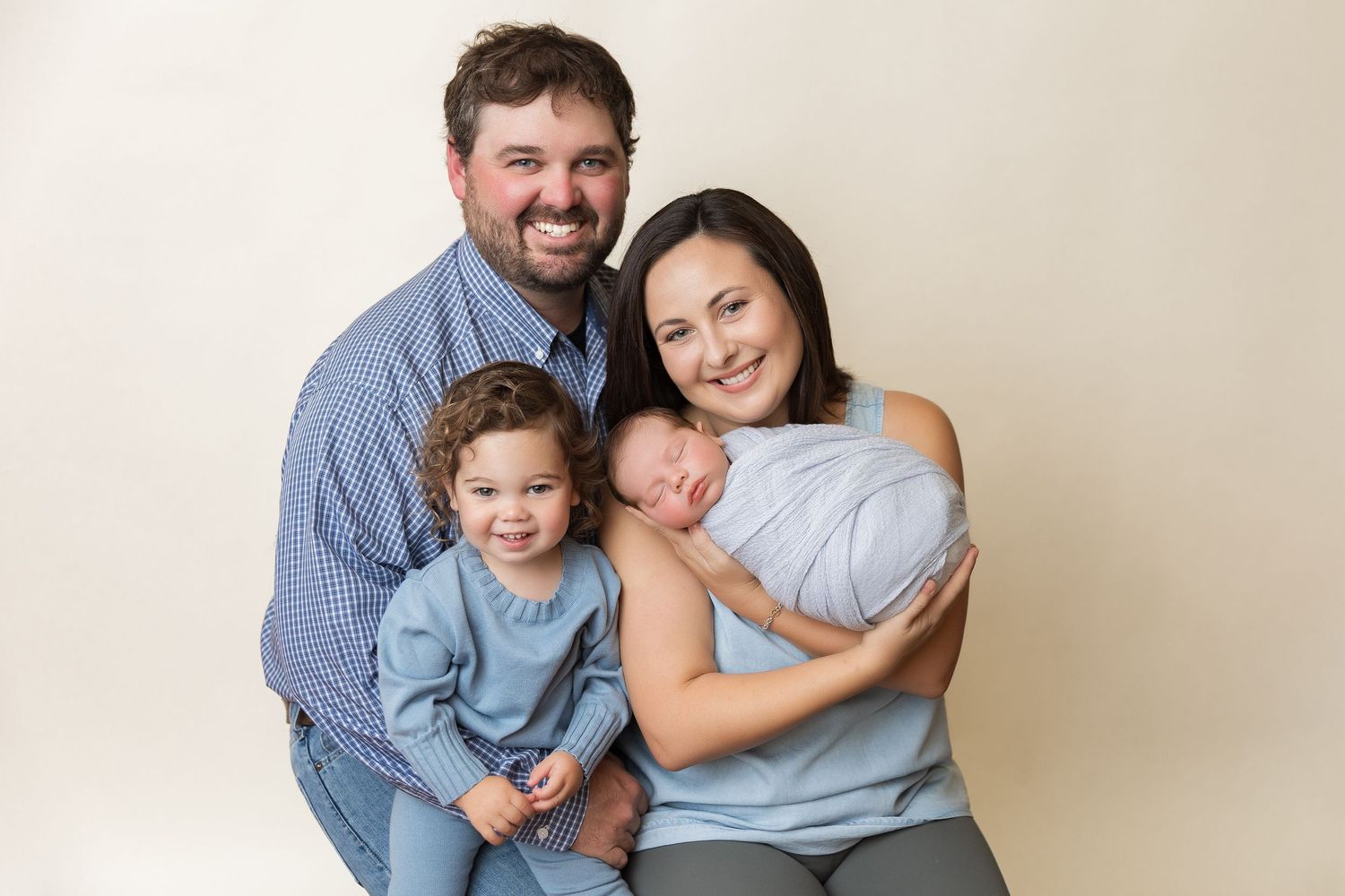Family portrait showing parents and baby in coordinating blue outfits against light background.