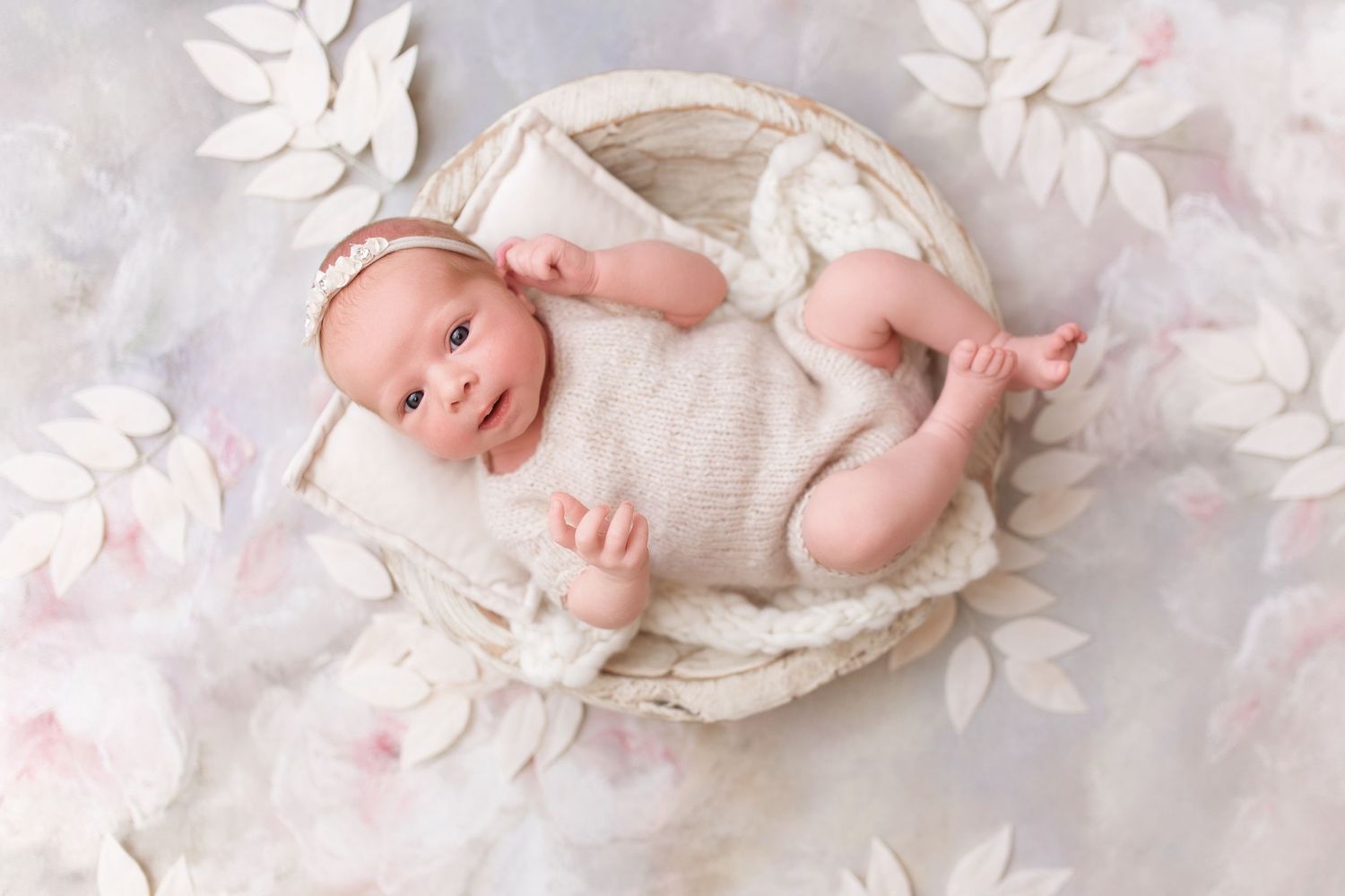 Newborn baby sleeping in white basket surrounded by delicate paper flowers.